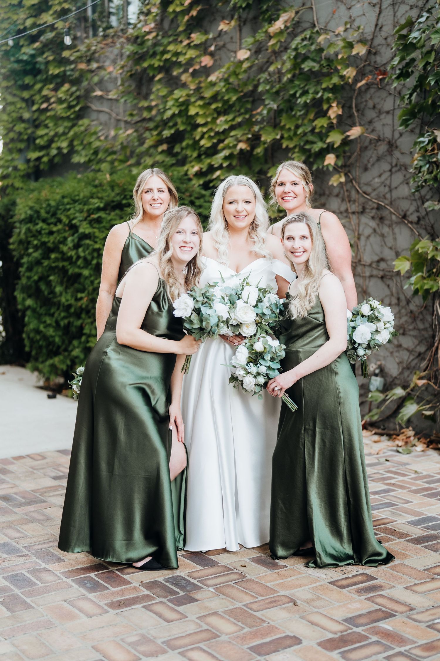 Wedding party members in green dresses and white gown share candid moments on brick patio.