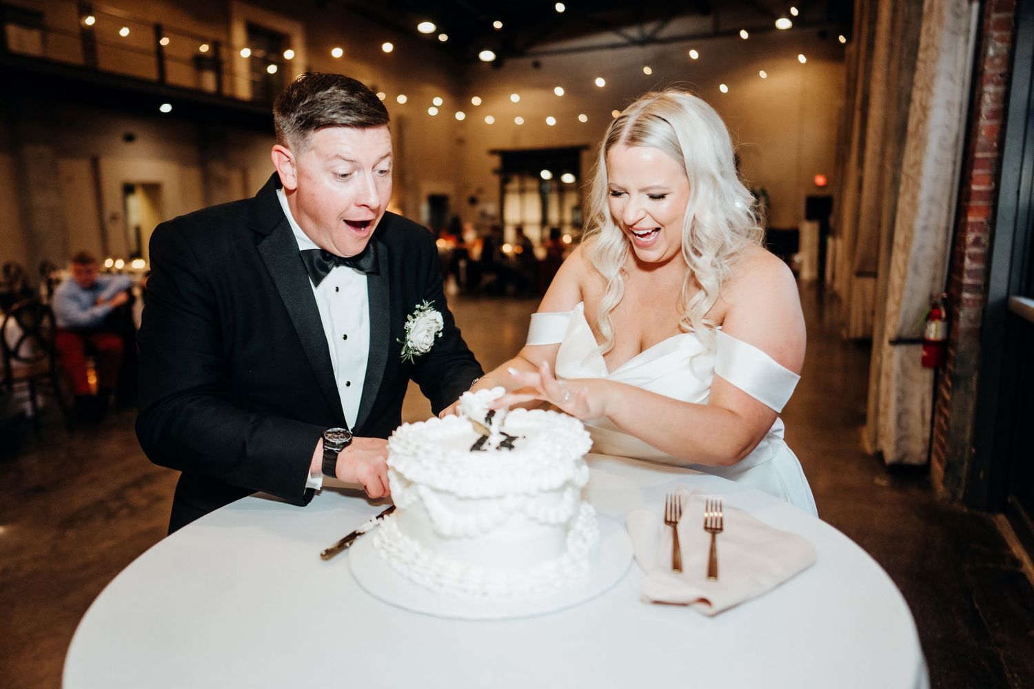 Couple shares cake cutting moment at wedding reception under warm string lights.