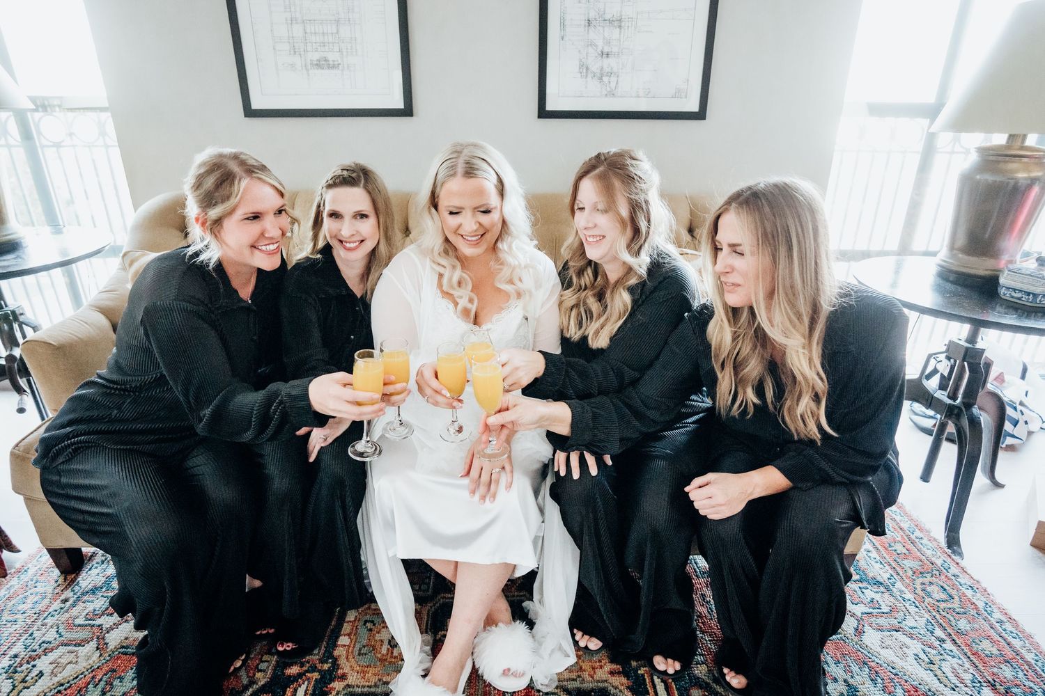 Bridesmaids in black dresses gather around the bride in a joyful celebration moment on a patterned carpet.