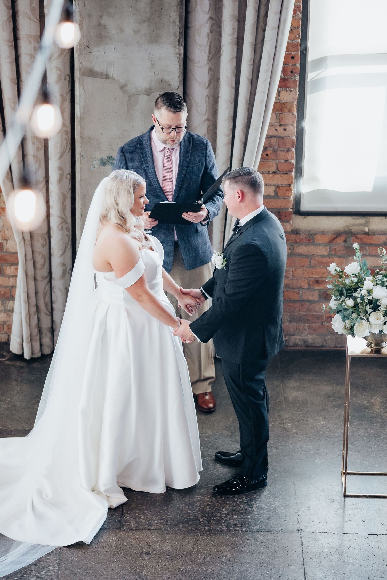 Wedding ceremony in an industrial brick venue with exposed beams and hanging lights during an intimate exchange of vows.