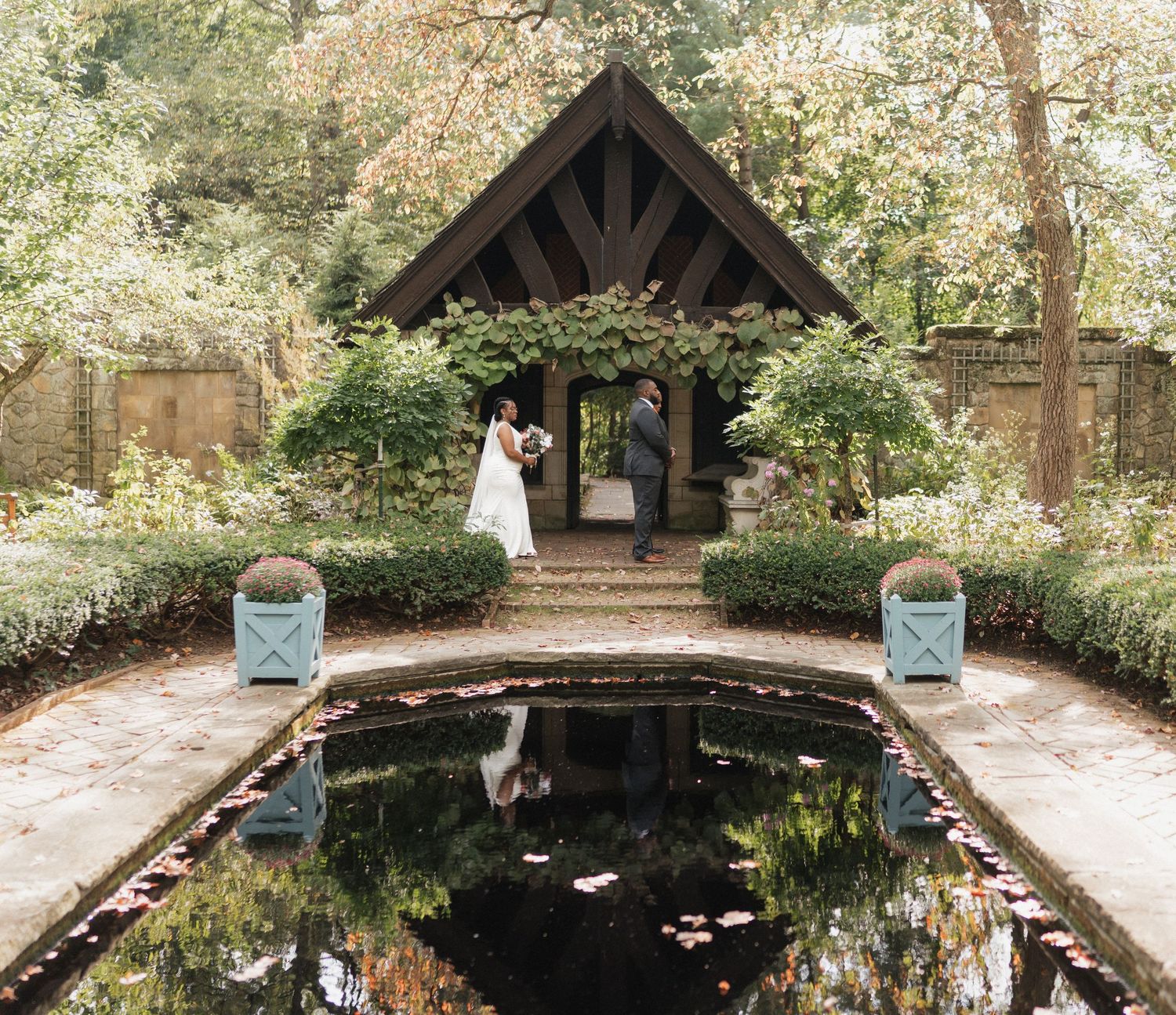 A rustic wooden pavilion nestled among trees and greenery with a tranquil pond in the foreground.