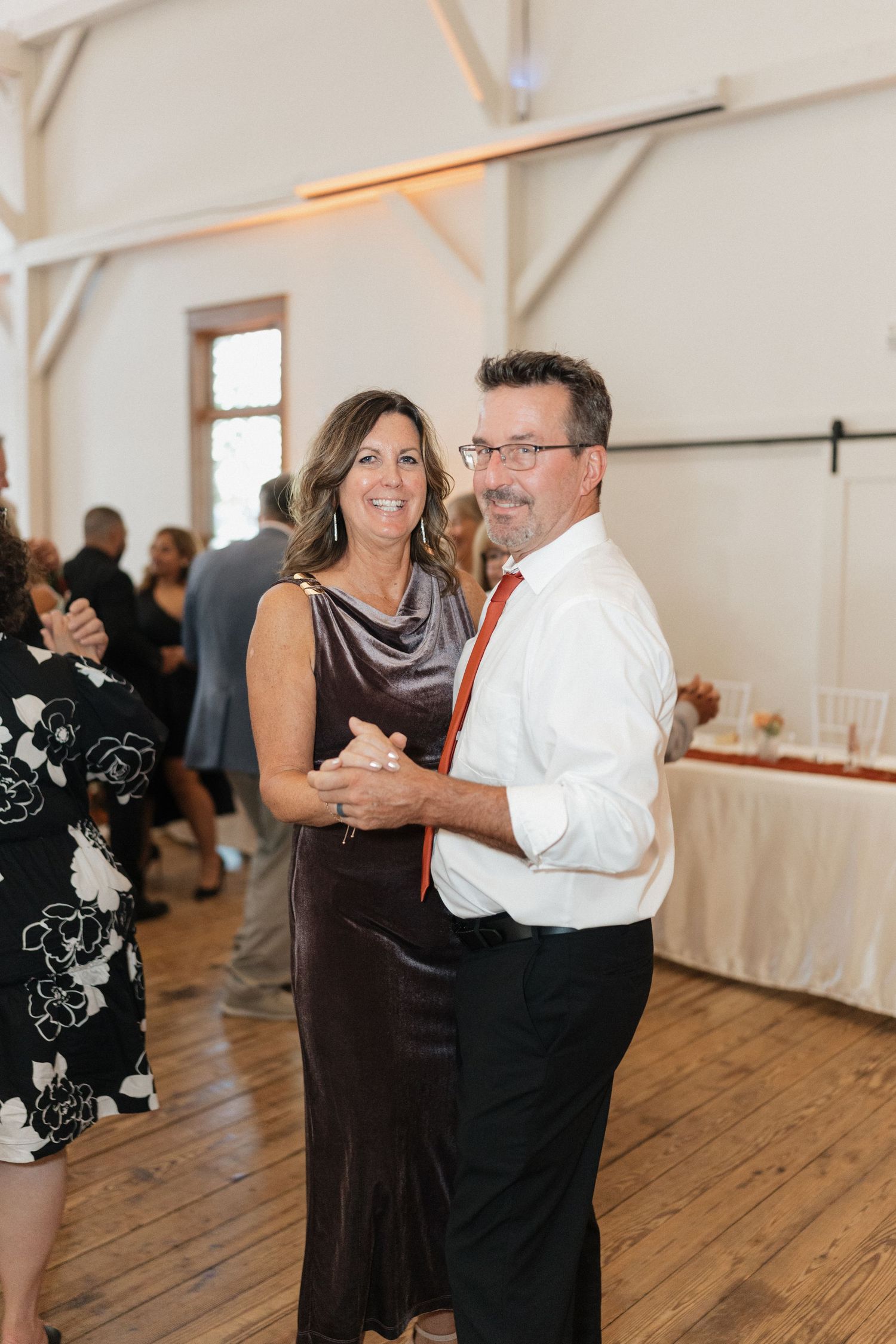 Wedding reception guests dancing on wooden floor in a bright white venue with vaulted ceilings.