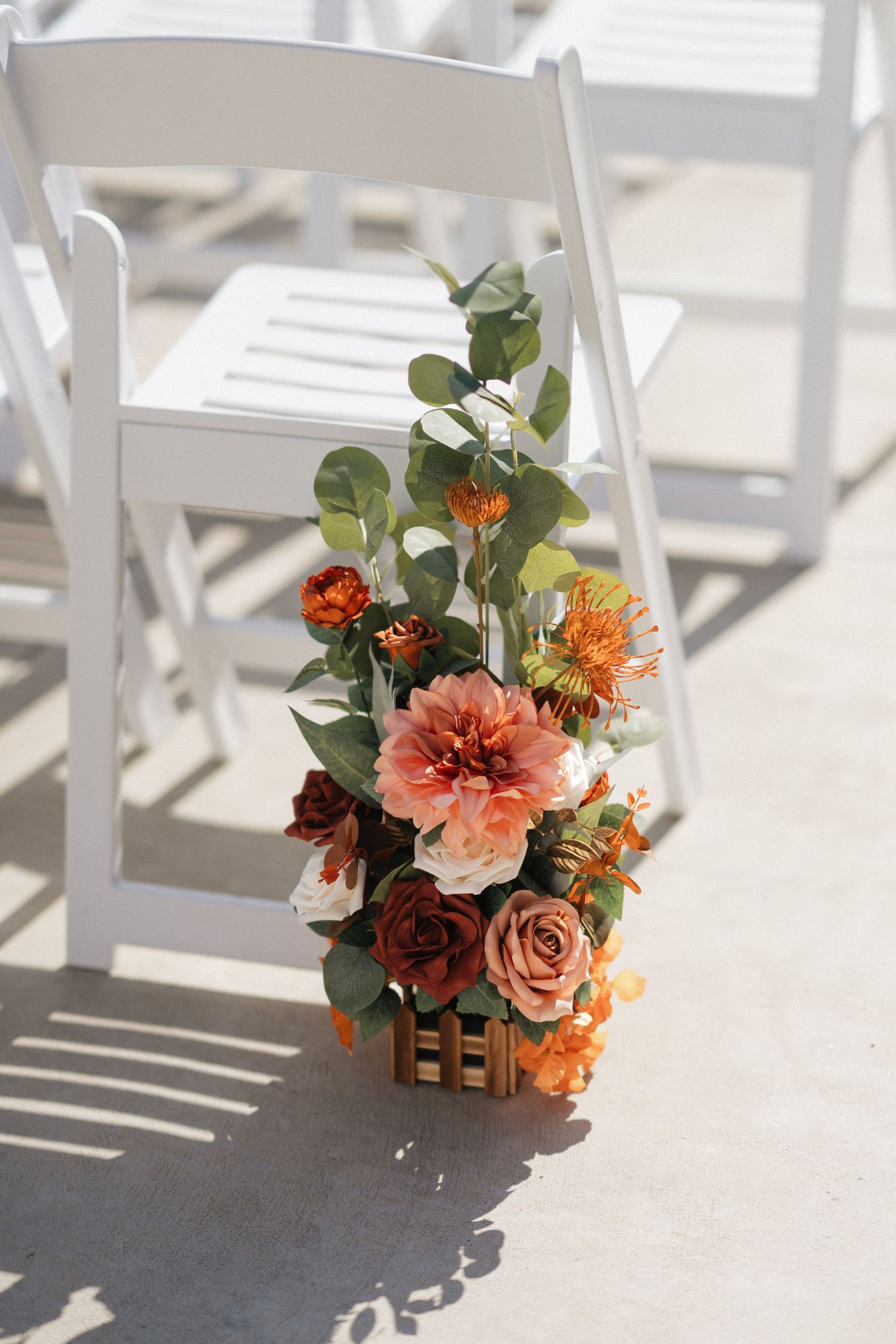 Orange and pink floral arrangement with eucalyptus decorates a white folding chair in natural sunlight.