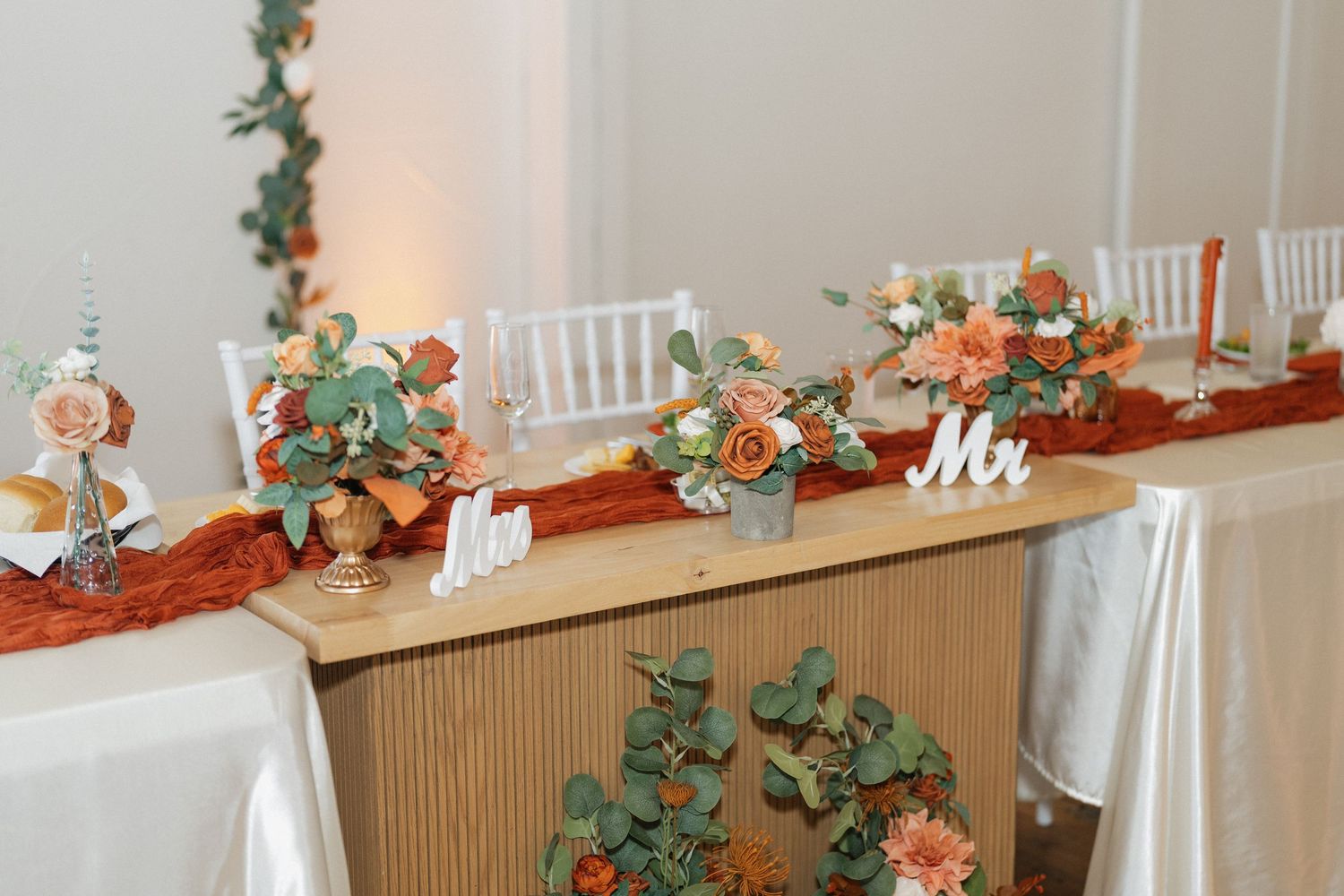 Wedding reception table decorated with greenery and orange floral arrangements.