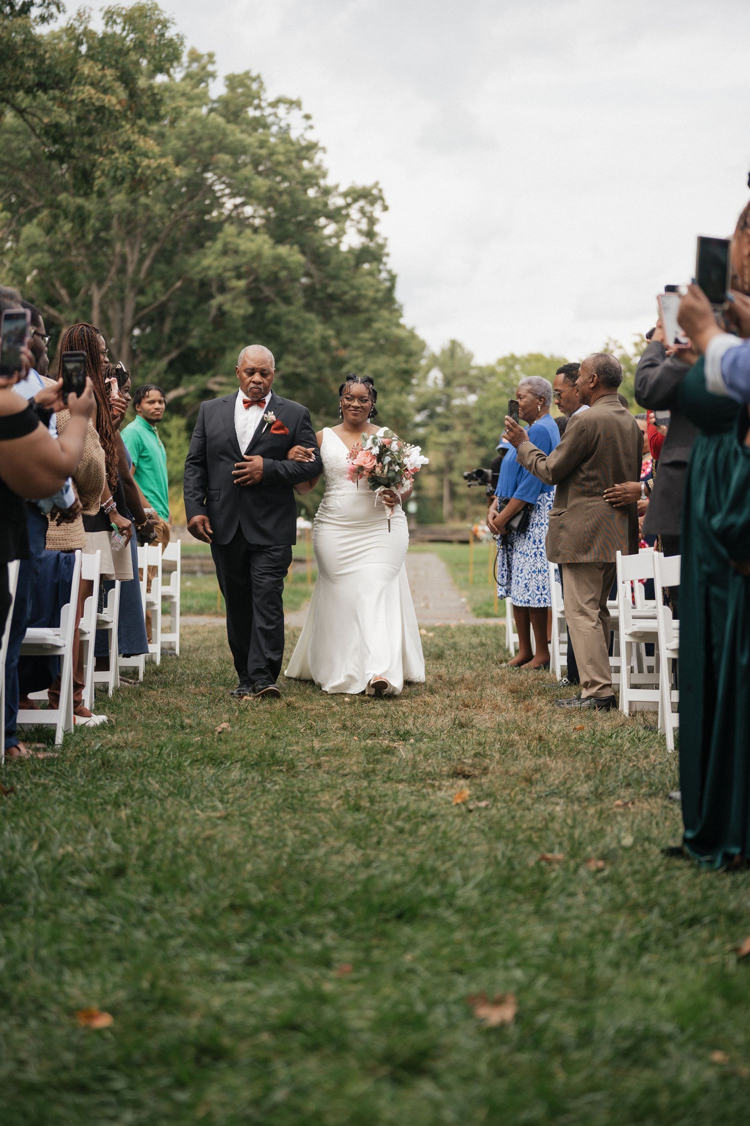 Wedding guests line up outdoors on a grassy path with white lanterns marking the aisle during an autumn ceremony.