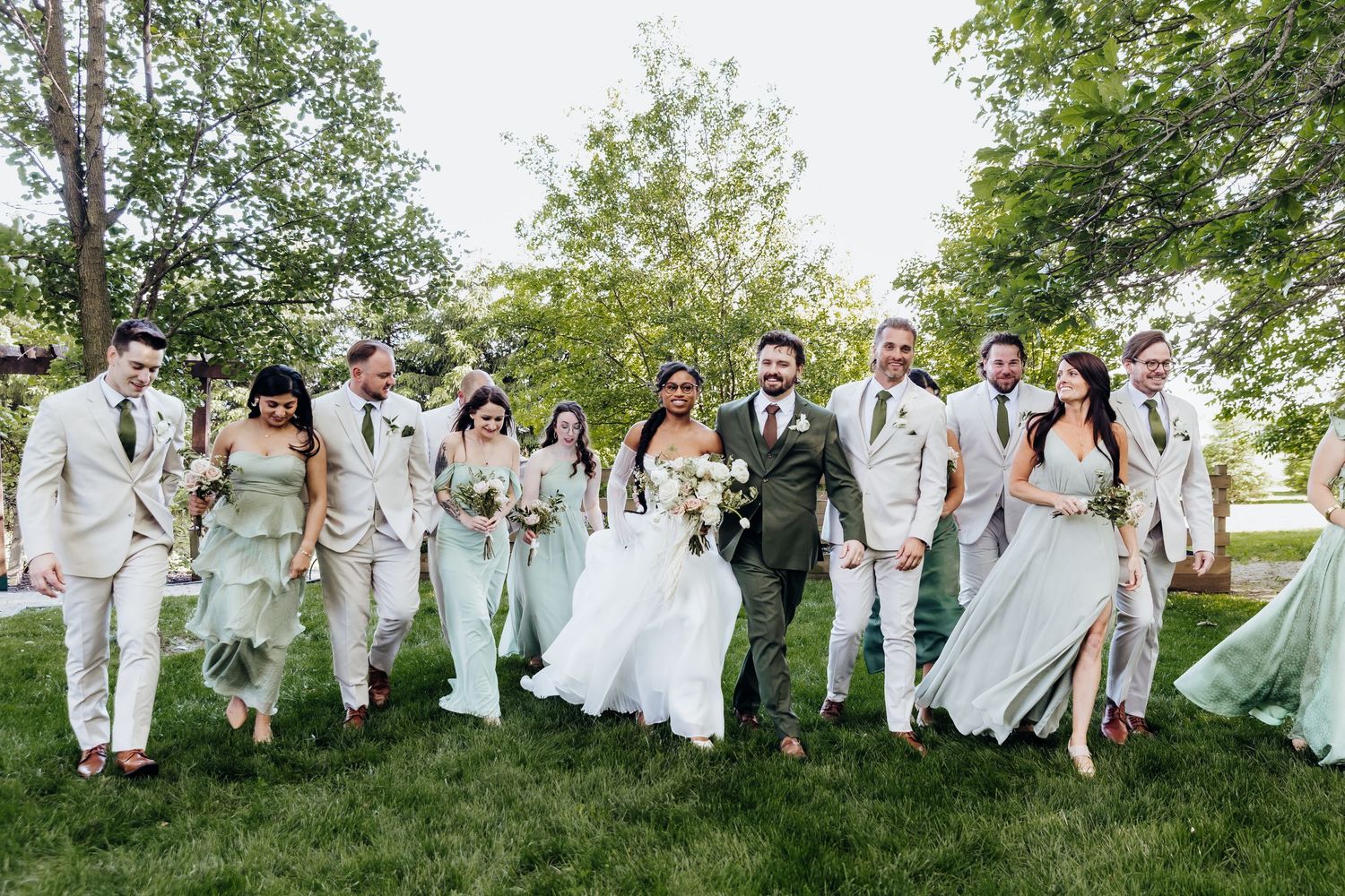 Wedding party in light colored attire walks together on grass during outdoor celebration.