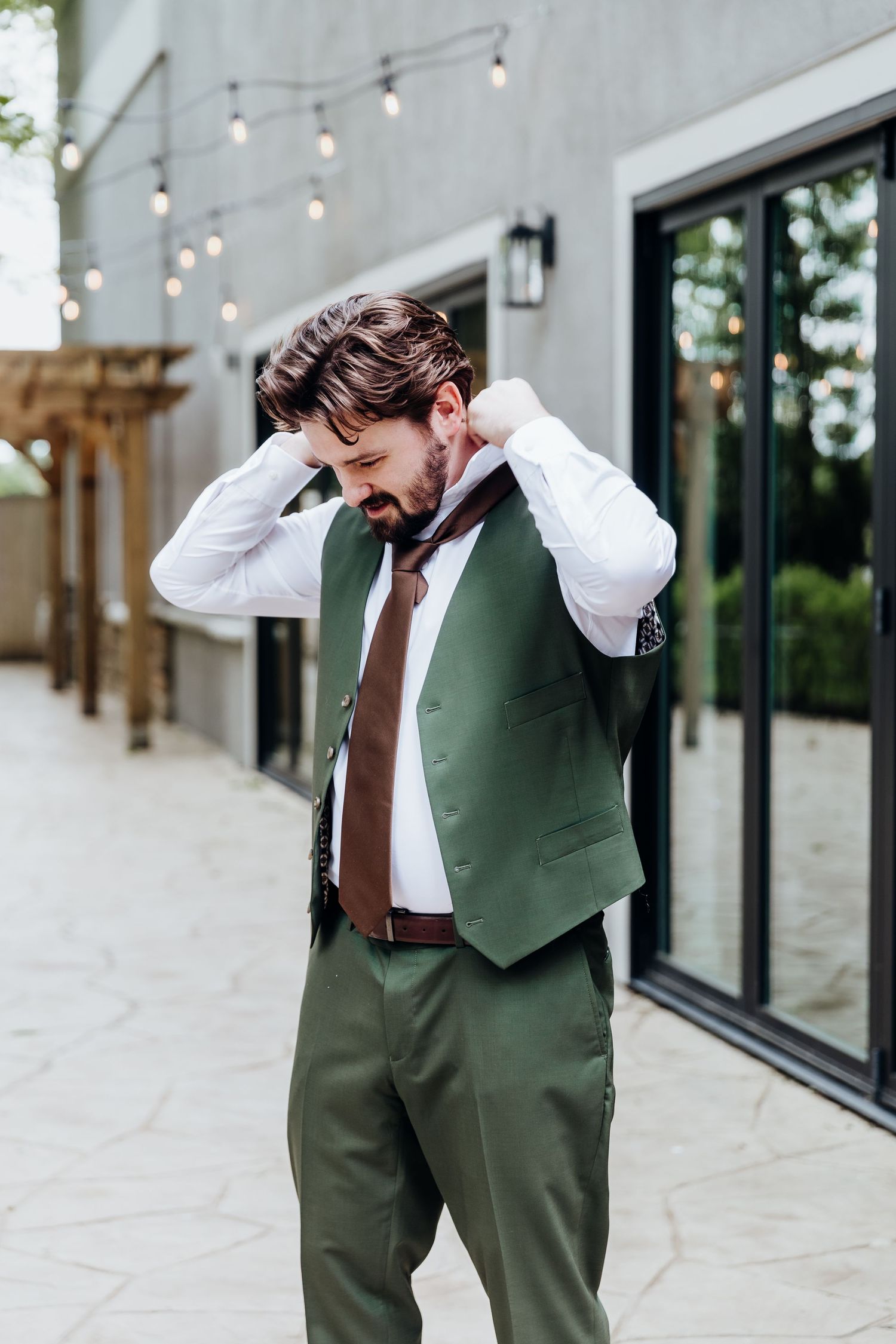 A groom in a green suit vest and brown tie adjusts his collar outdoors under string lights.