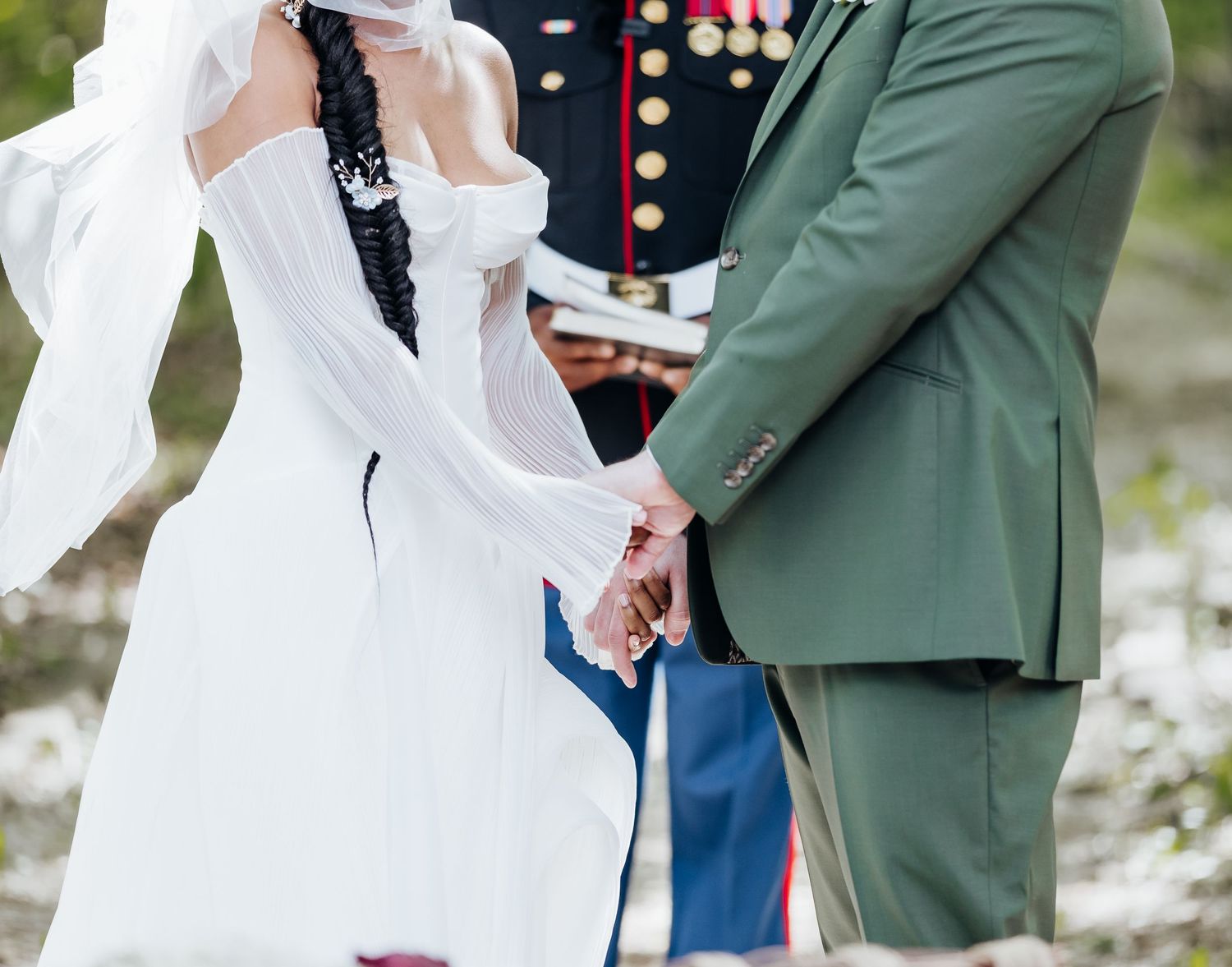 Wedding couple in green suit and white dress hold hands during outdoor ceremony.