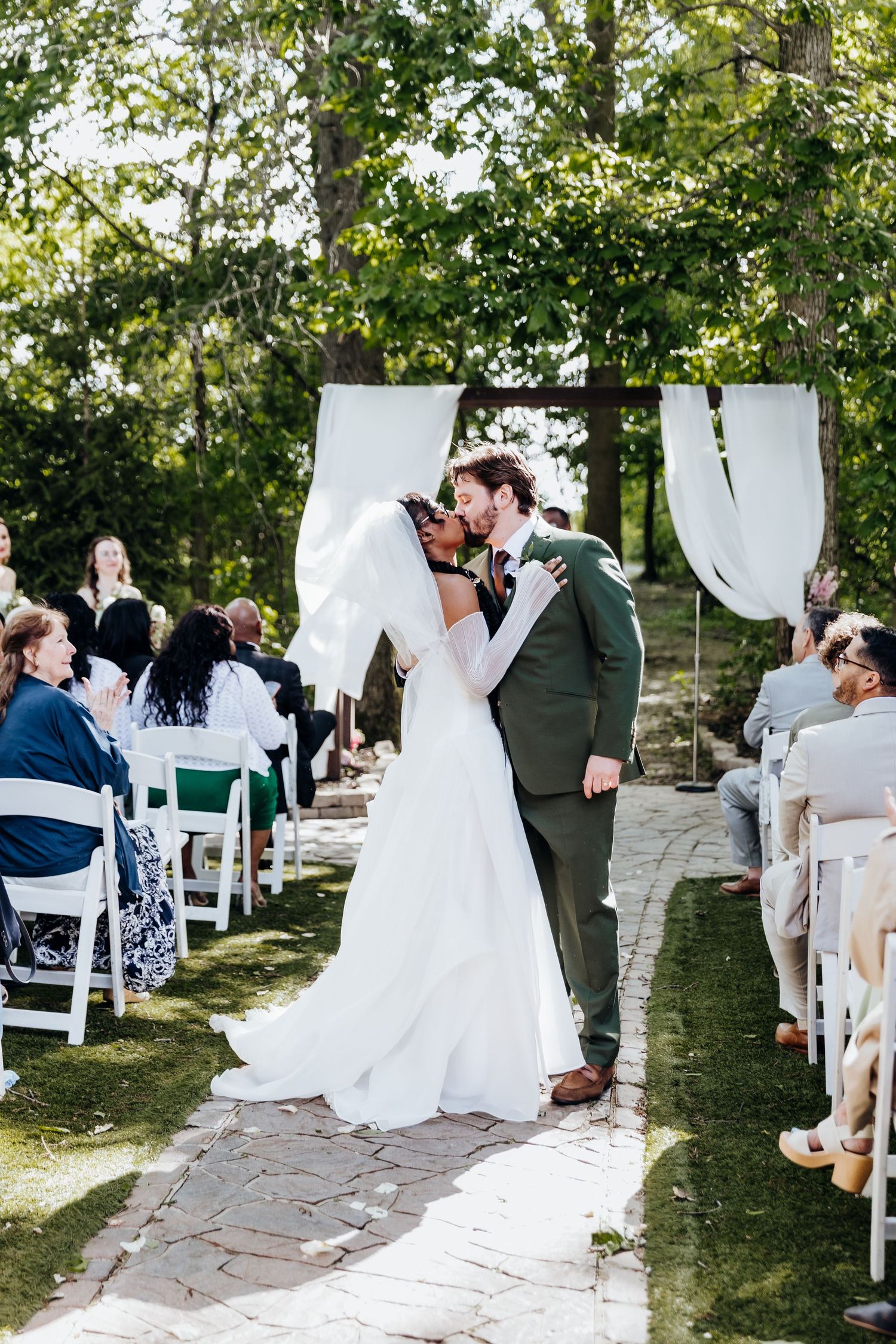 Wedding ceremony moment under white fabric arch with guests seated on white chairs.