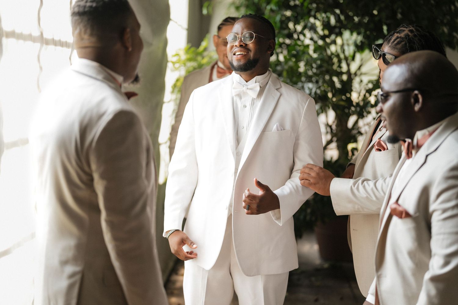 Wedding party members in white suits laugh together during pre-ceremony preparations.