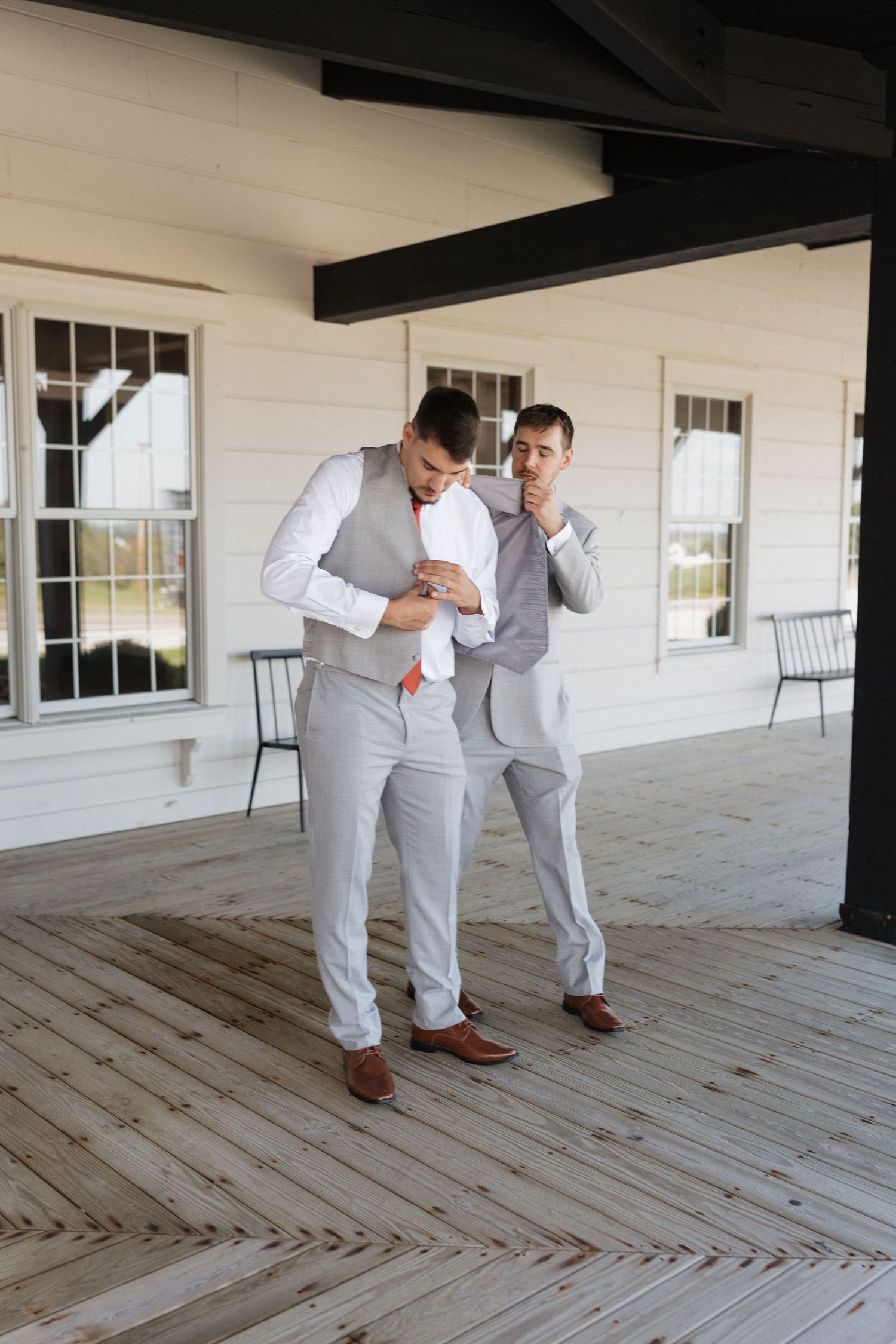 Two groomsmen in light gray suits share a playful moment on a wooden porch during a wedding photoshoot.