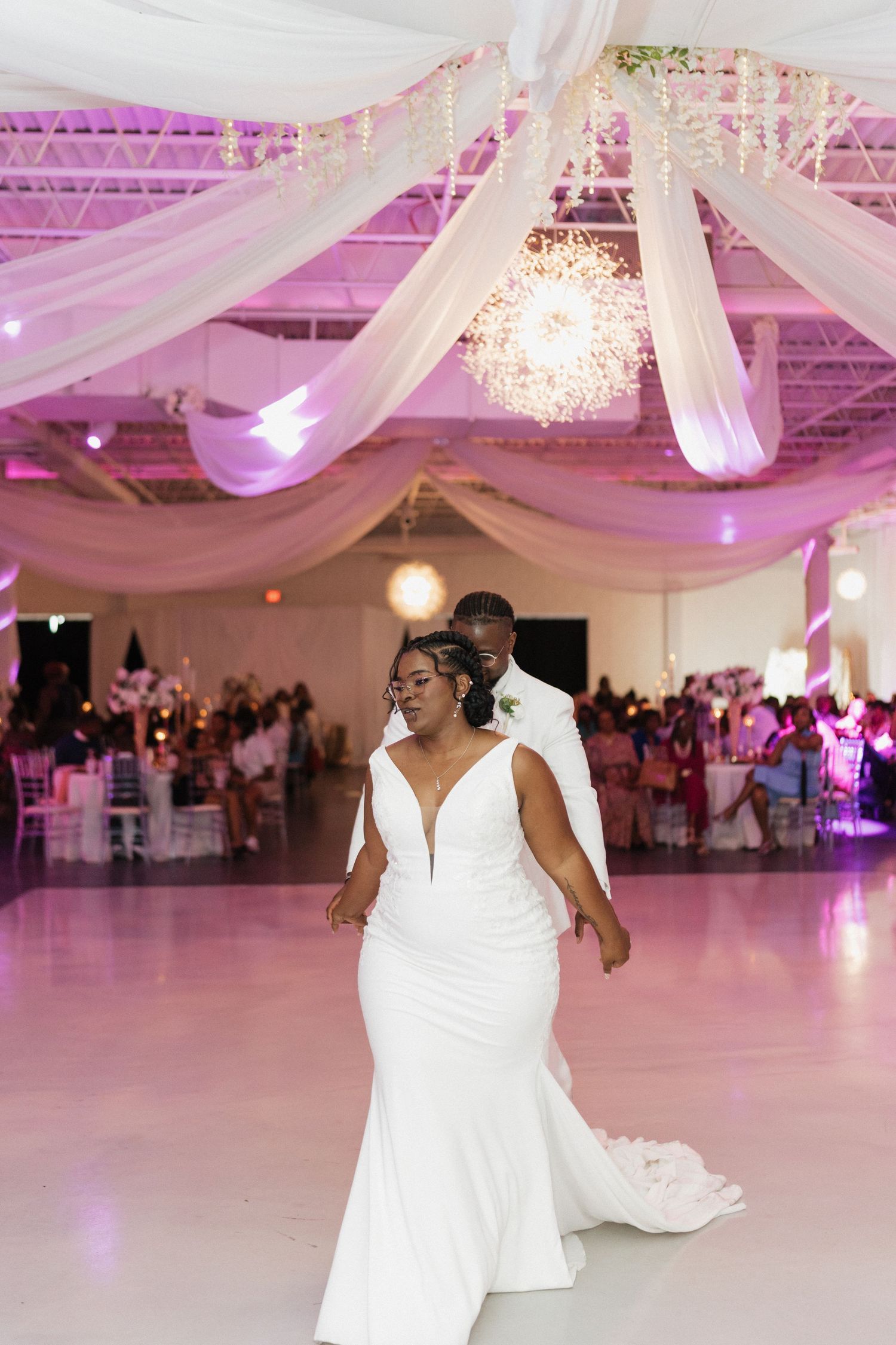Wedding reception venue decorated with white draping and pink lighting featuring couple dancing.