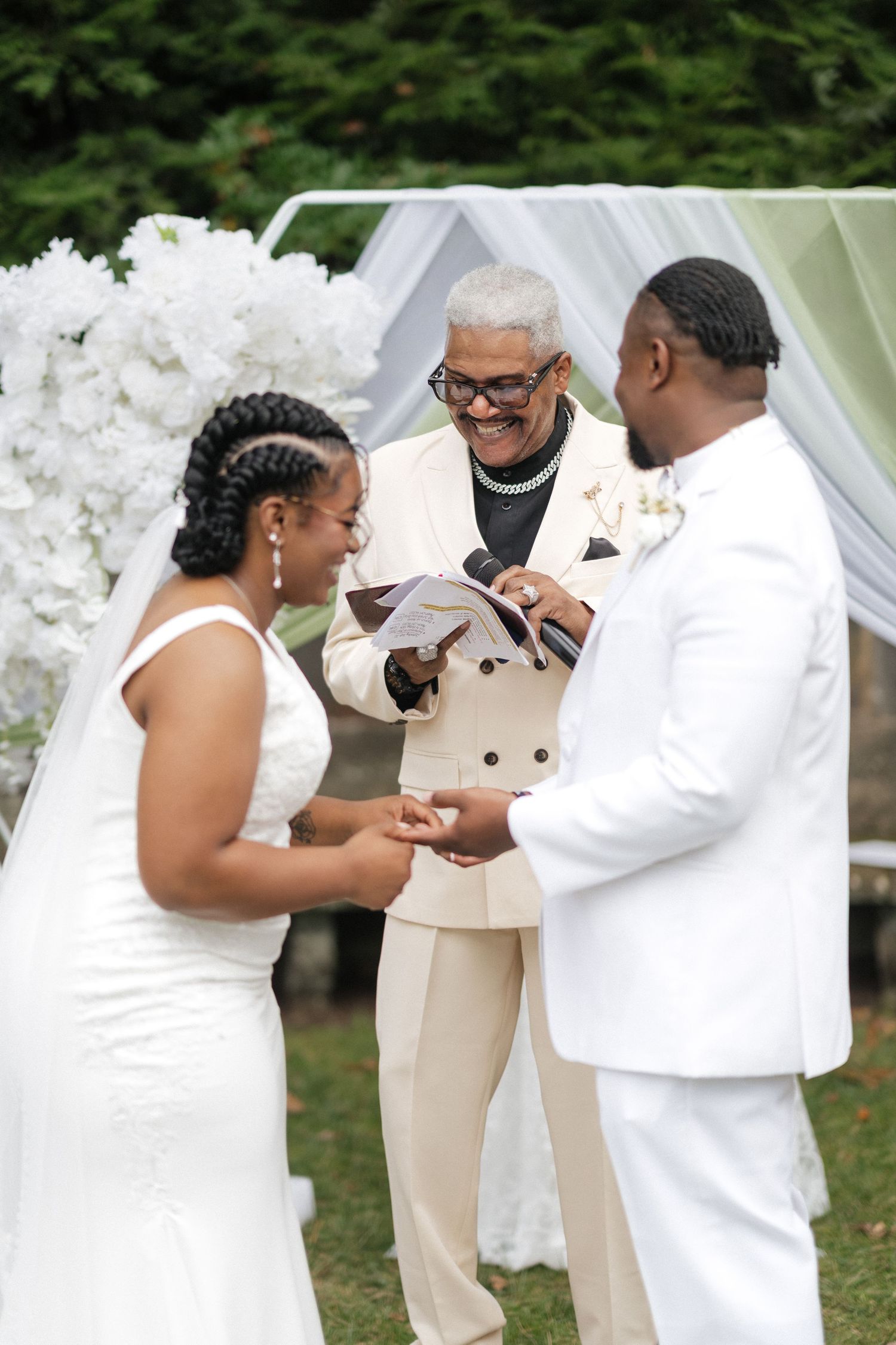 A wedding ceremony takes place outdoors with an officiant in beige and couple in white exchanging rings under a white canopy.