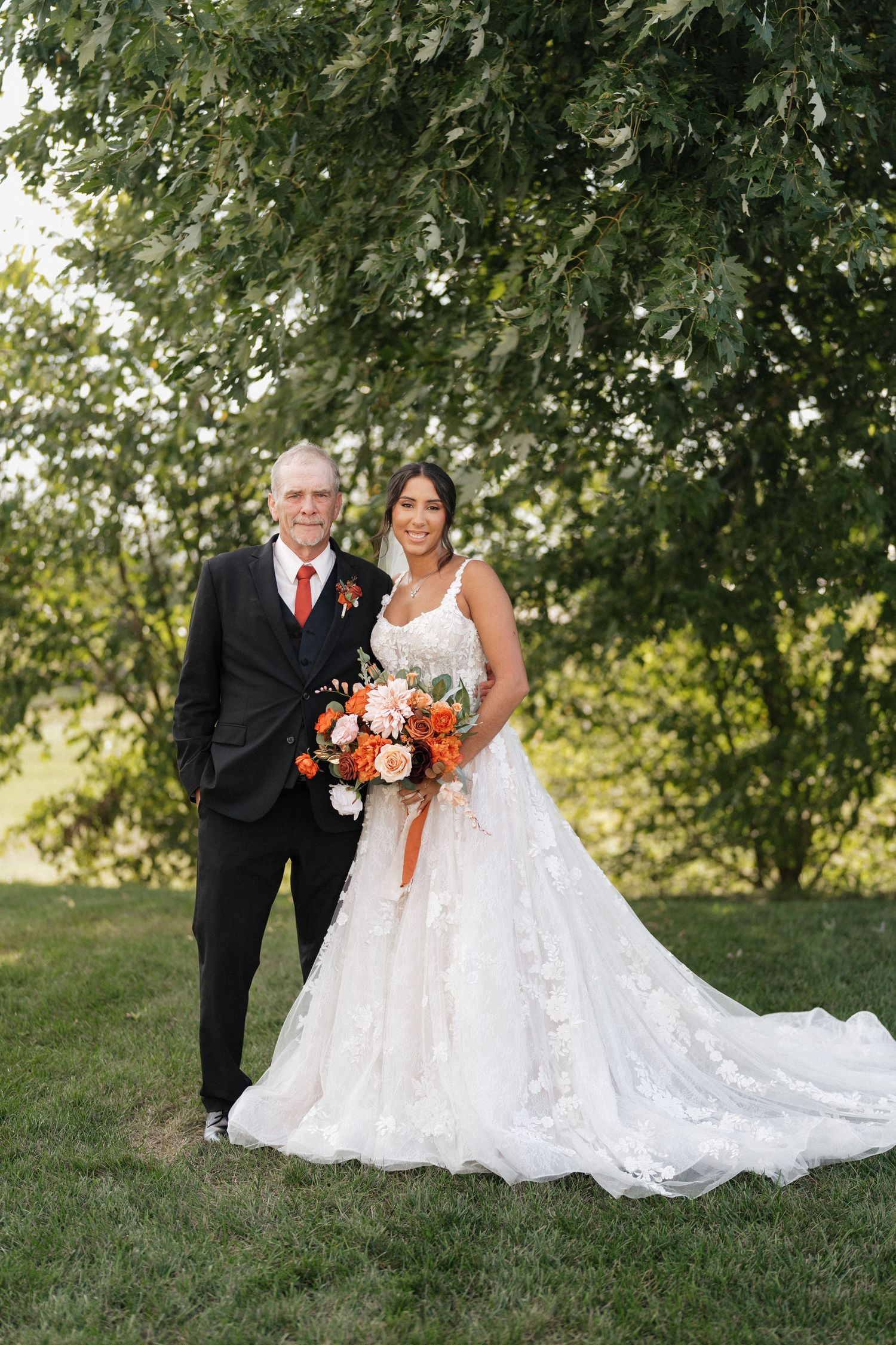 A couple poses outdoors in wedding attire with a flowing white dress and orange bouquet against a natural backdrop.