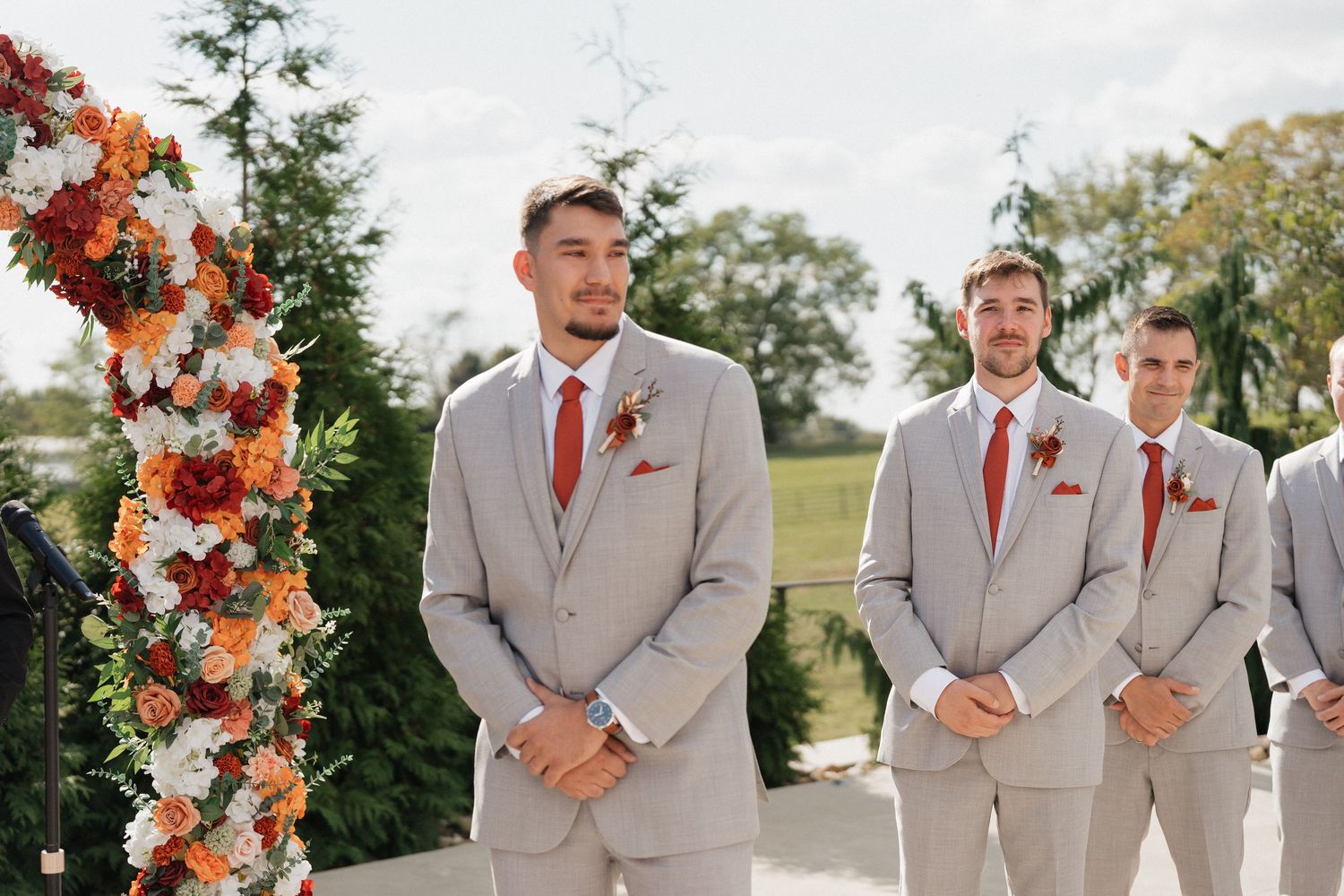 Wedding party in gray suits and rust-colored dresses pose by floral arch outdoors.