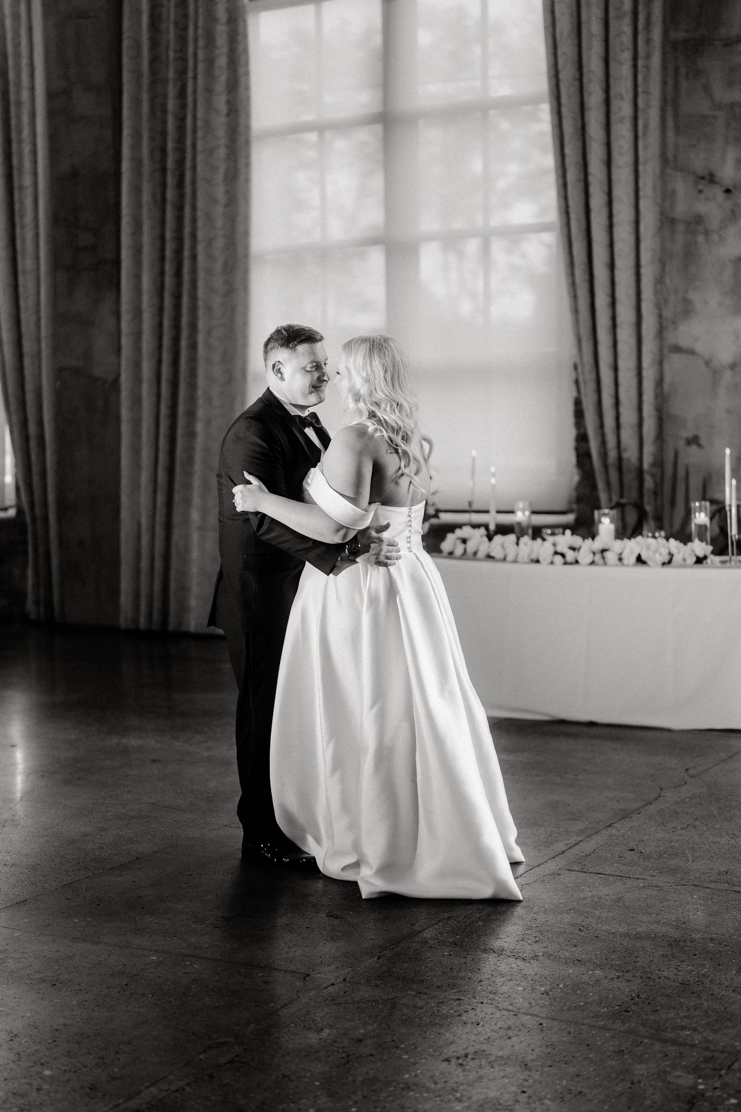 Black and white photo of couple dancing in elegant ballroom with dramatic window lighting.