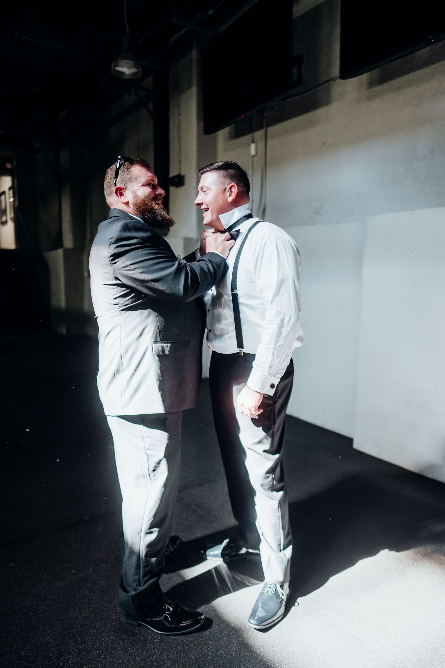 Two people in contrasting outfits adjusting formal wear in a dimly lit space.