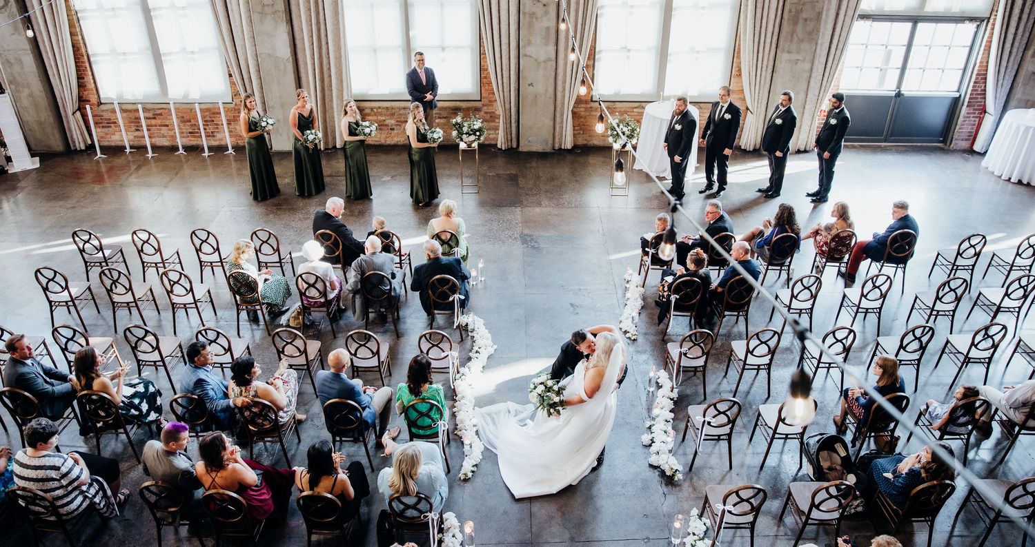 Wide angle view of a wedding ceremony setup in an airy industrial venue with vintage chairs.
