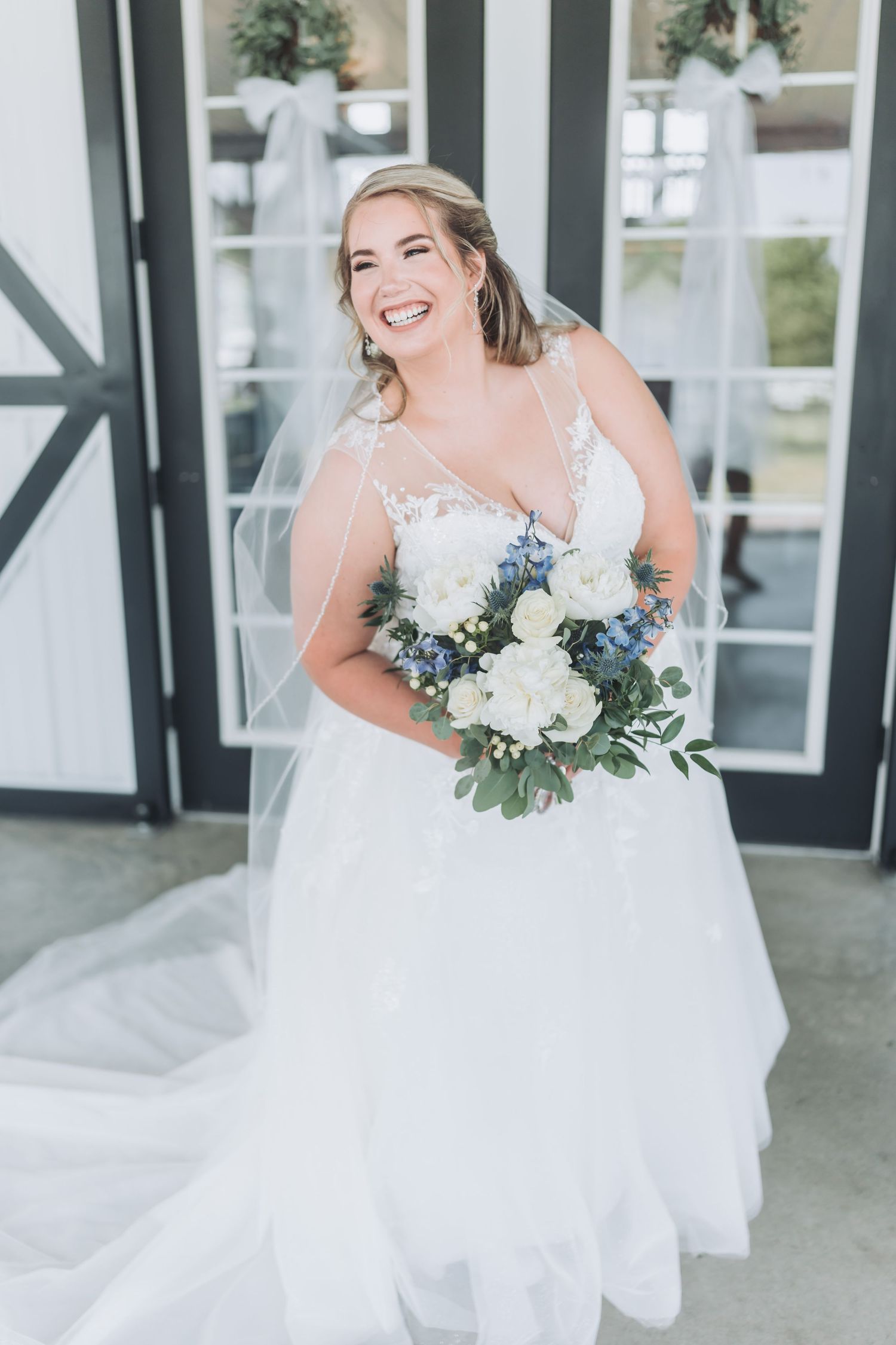 Radiant bride in flowing white gown holds bouquet of white flowers outside rustic venue.