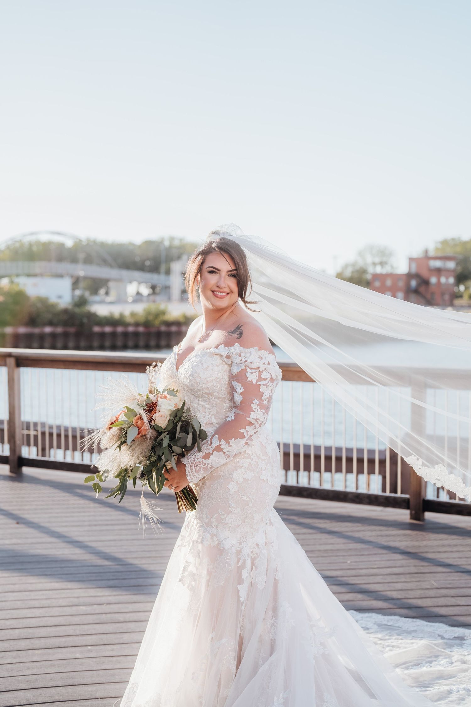 Bride in lace gown with flowing veil poses on waterfront deck holding rustic bouquet.
