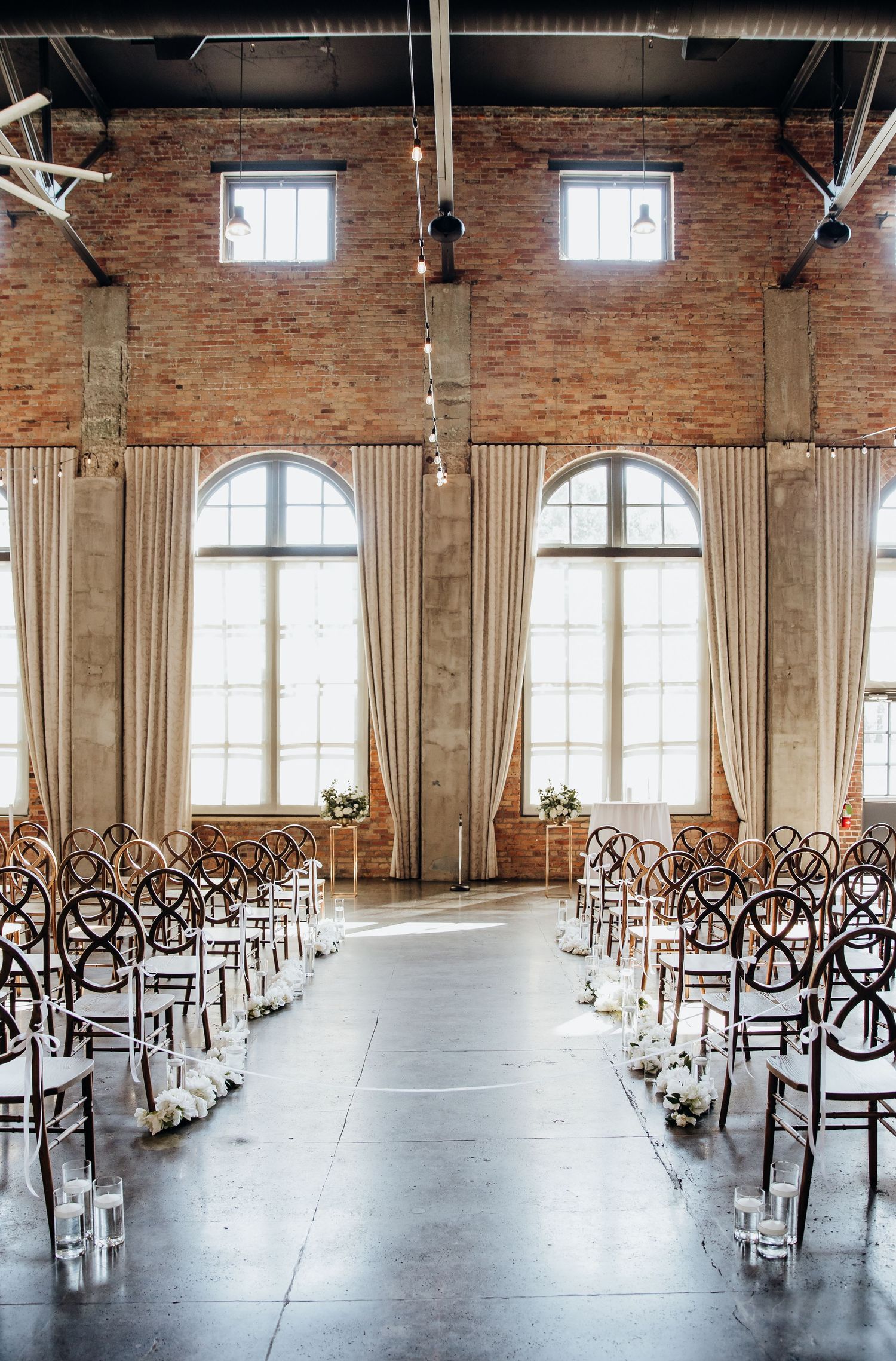 Empty wedding ceremony setup with cross-back chairs in an industrial brick venue with tall windows.