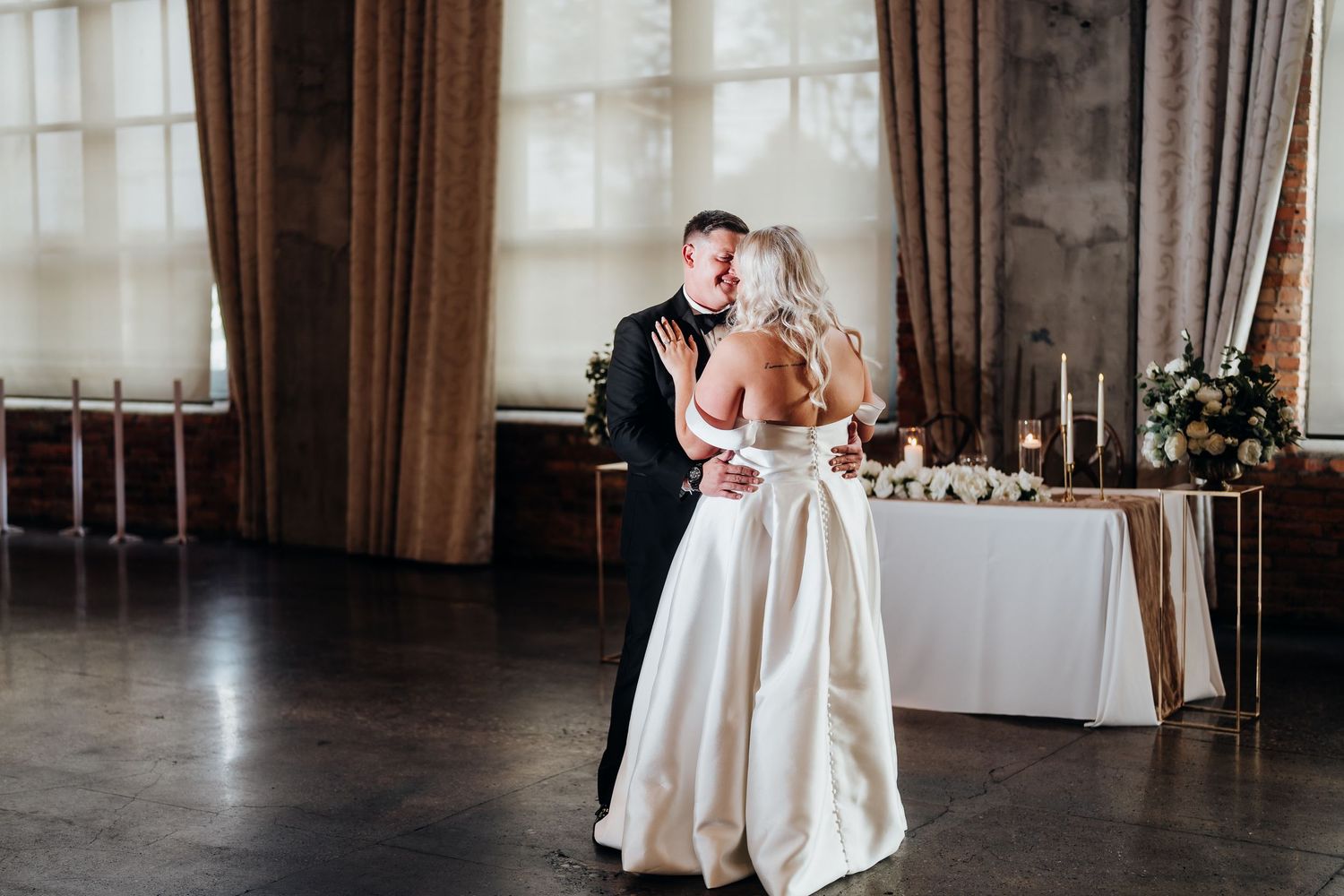 A couple dances together in an elegant ballroom with large windows and draped curtains.