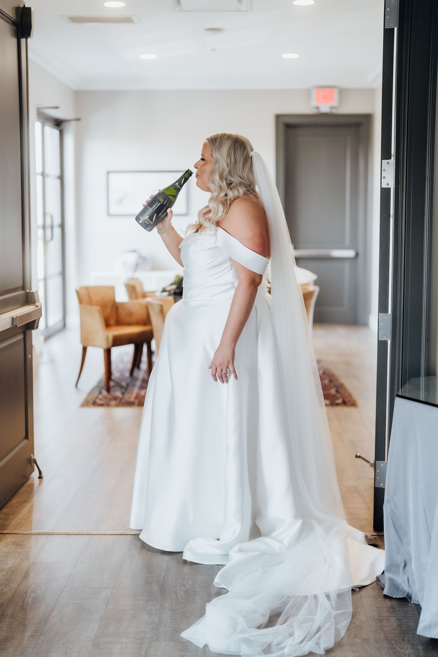 A wedding dress train trails elegantly across a bright hallway floor with white walls and wooden accents.