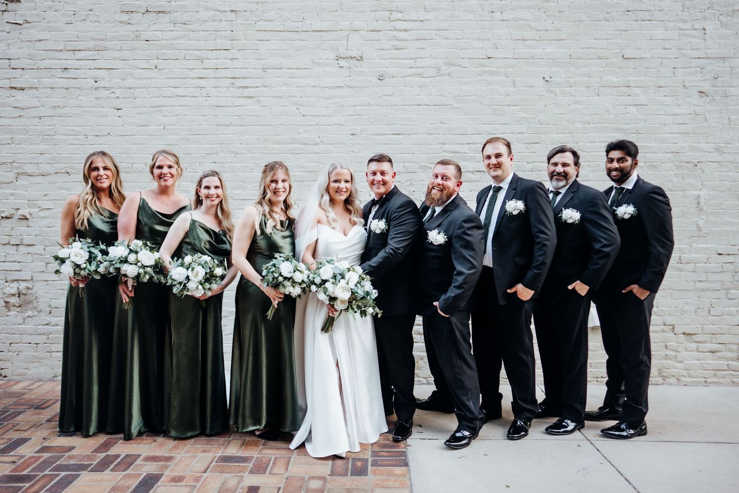 Wedding party poses against white brick wall wearing black suits and dark green dresses.