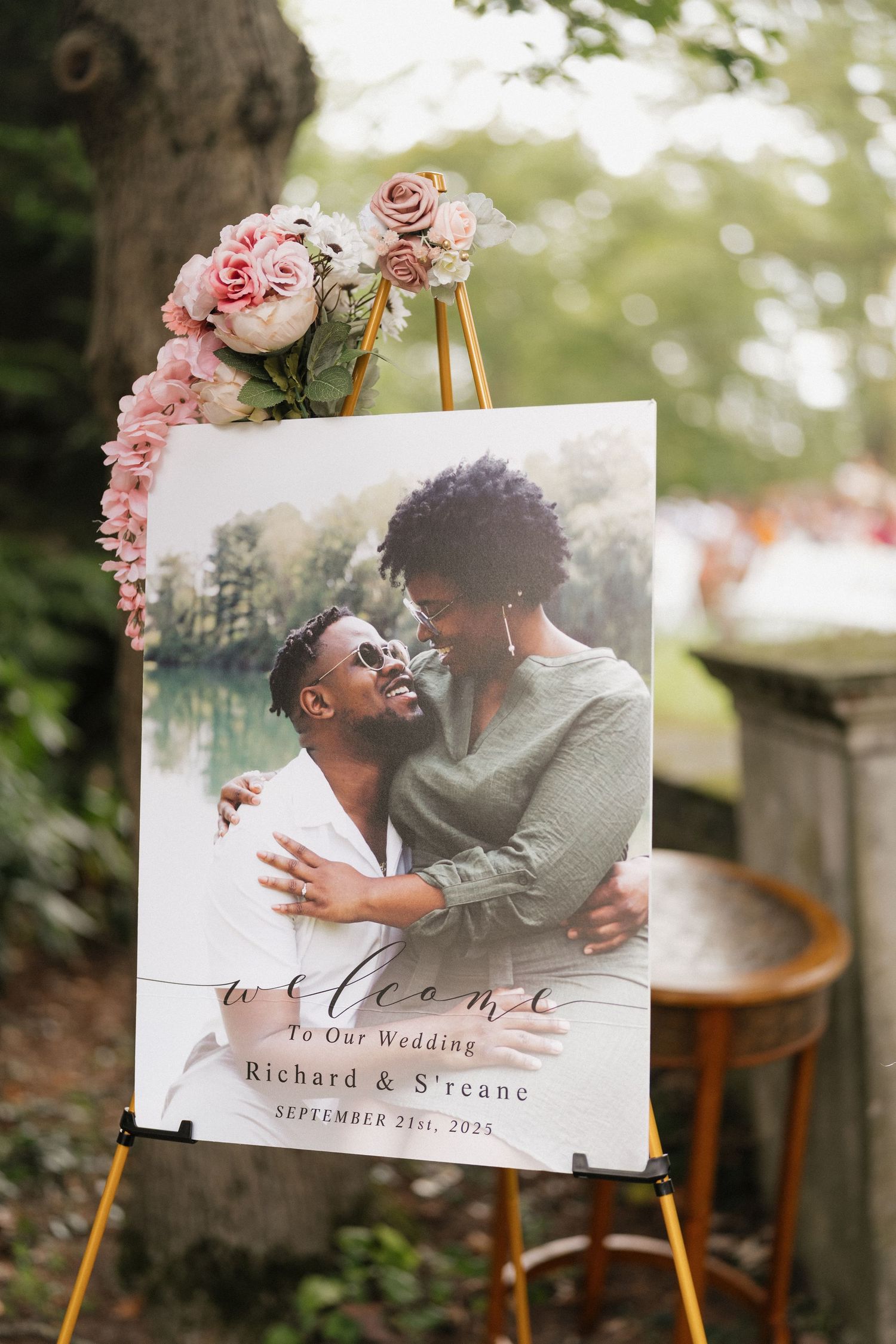 Wedding welcome sign with pink roses and romantic couple photo displayed on wooden easel in outdoor garden setting.