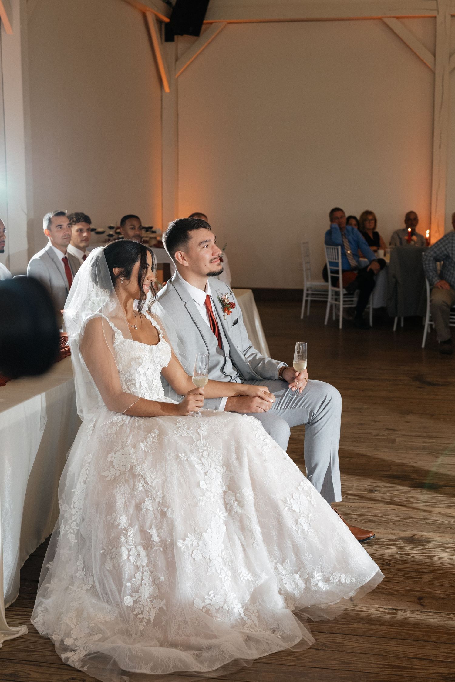 Wedding couple and guests seated at reception tables in a rustic indoor venue with hardwood floors.