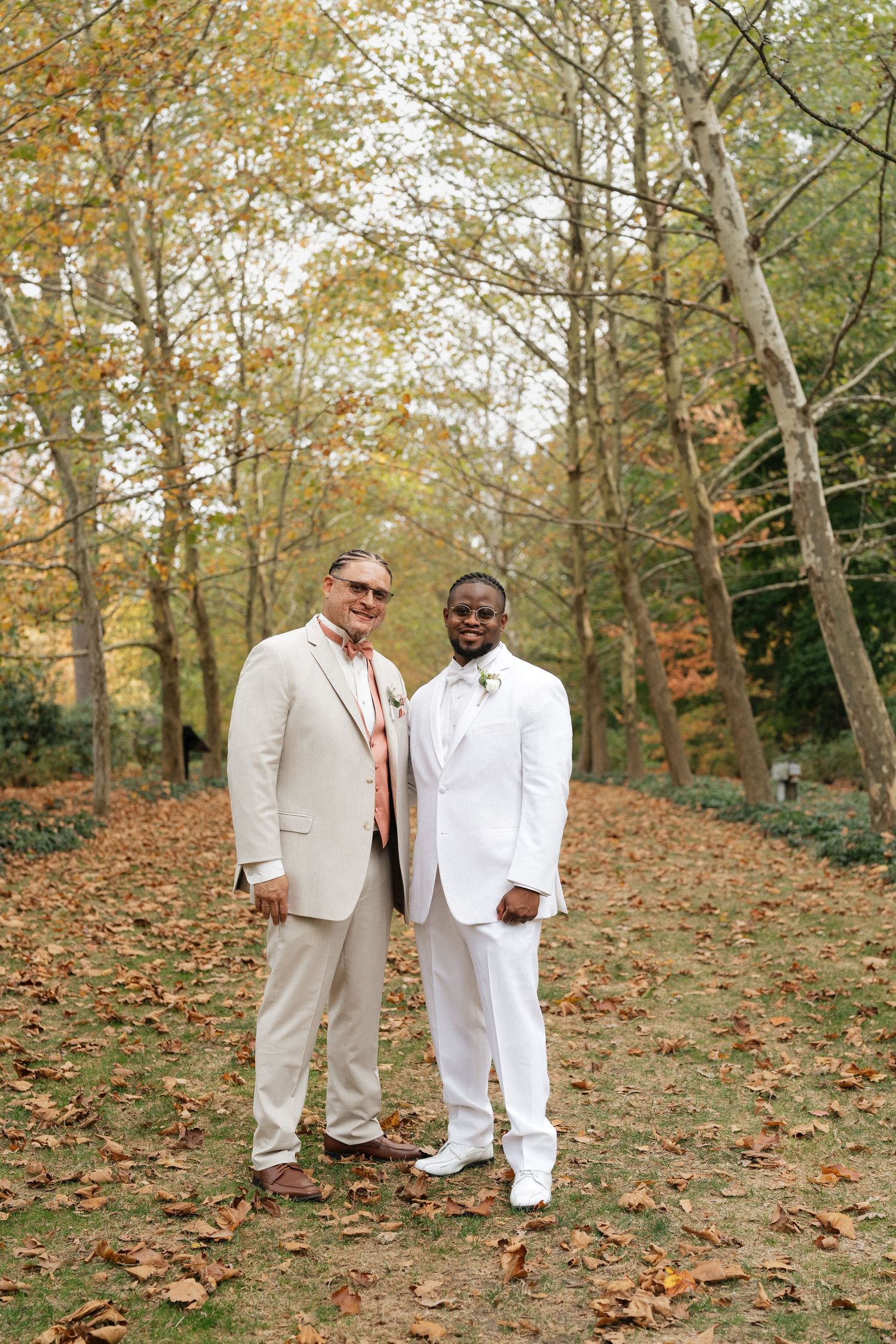 Two people in white suits pose among autumn leaves in a wooded setting.