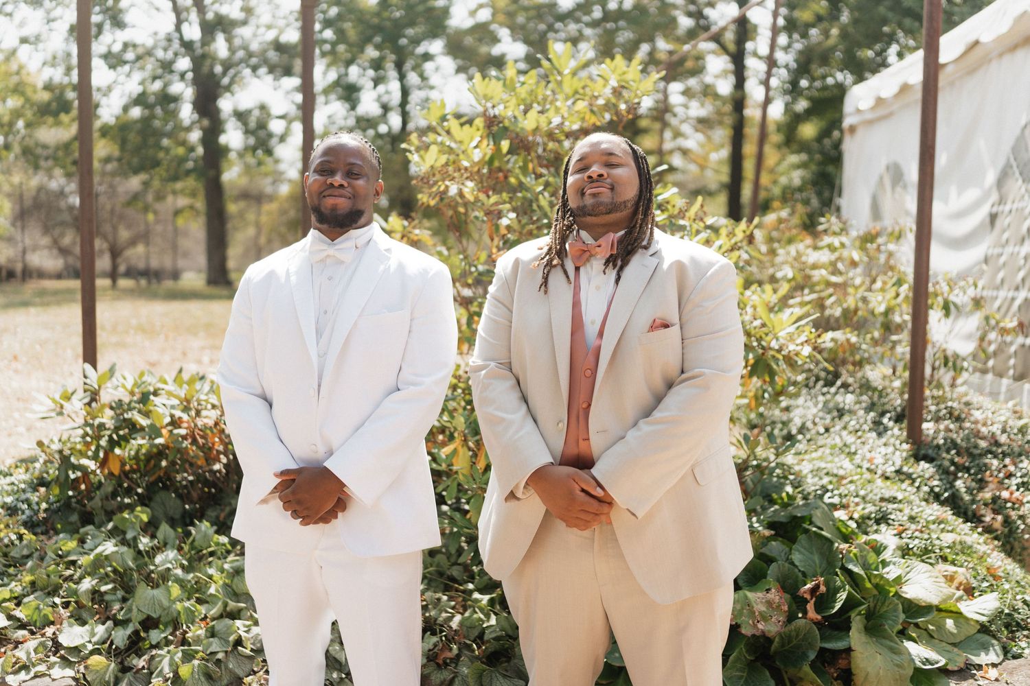 Two groomsmen in elegant white suits pose together in a garden setting.