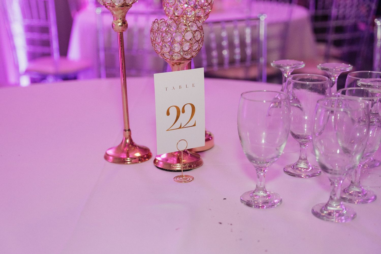 Wedding reception table detail with gold table number 22 and crystal glassware in purple lighting.