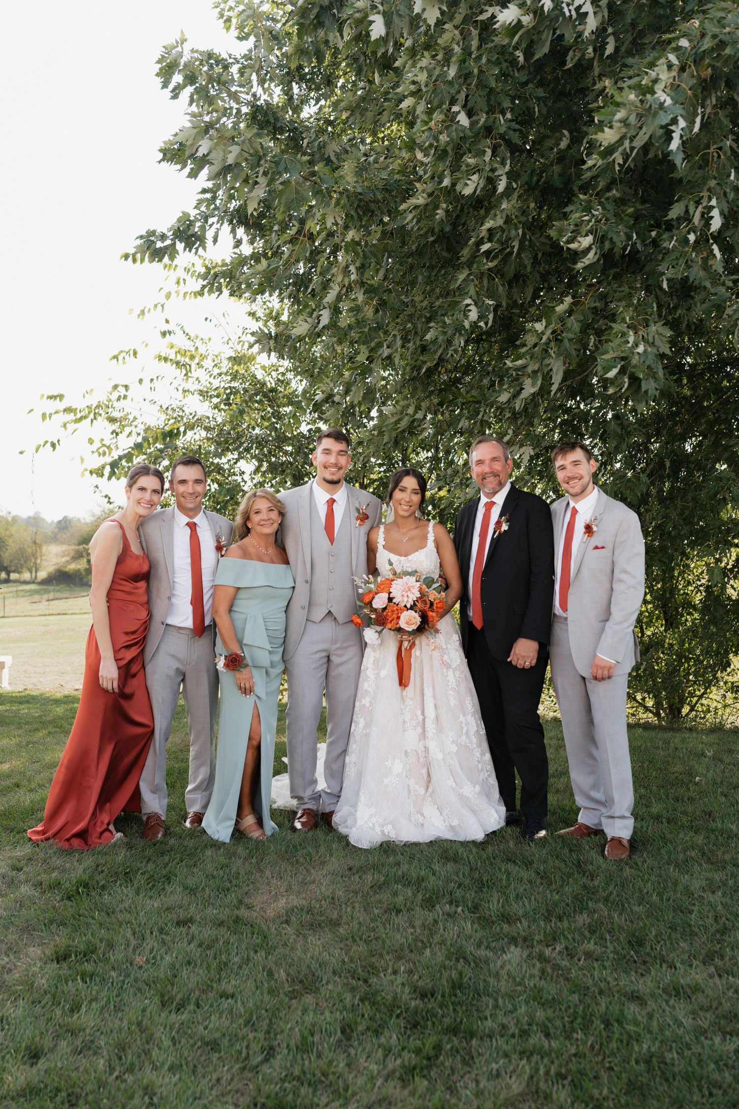 Wedding party in grey suits and rust-colored dresses posing together on grassy hill.