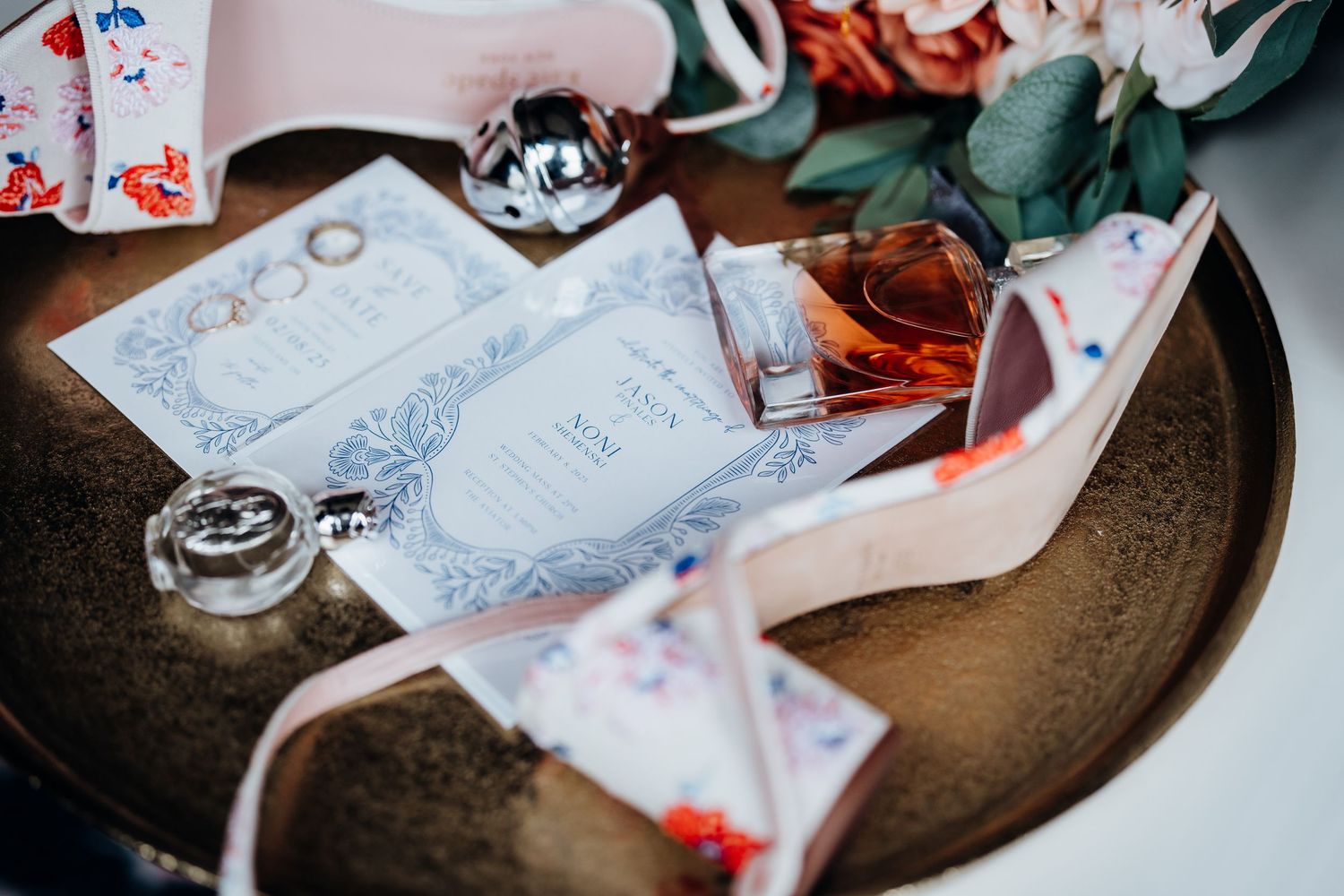 Wedding accessories including elegant white heels, blue invitation cards and decorative details arranged on a metallic plate.