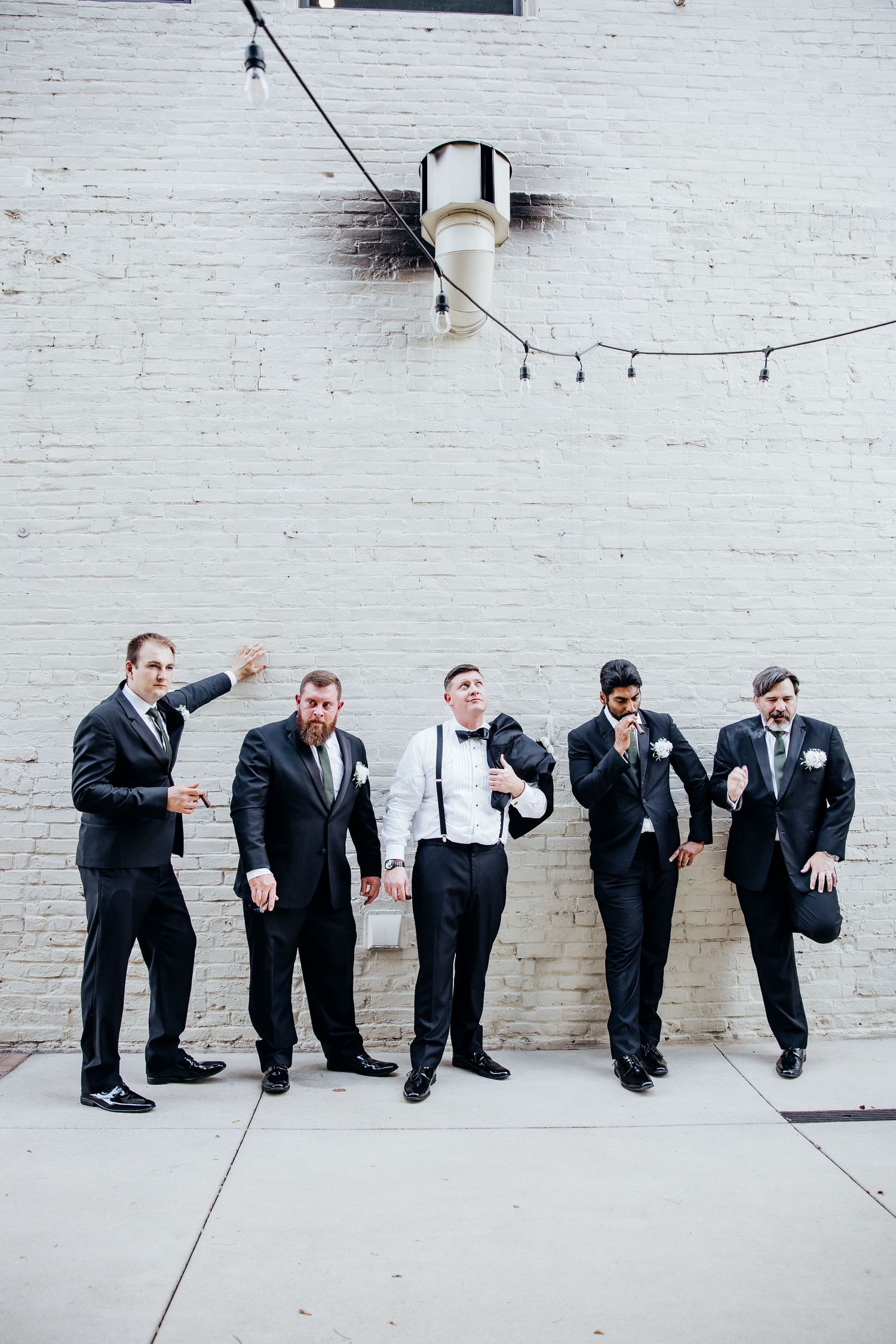 Groomsmen in black suits strike playful poses against a white brick wall.