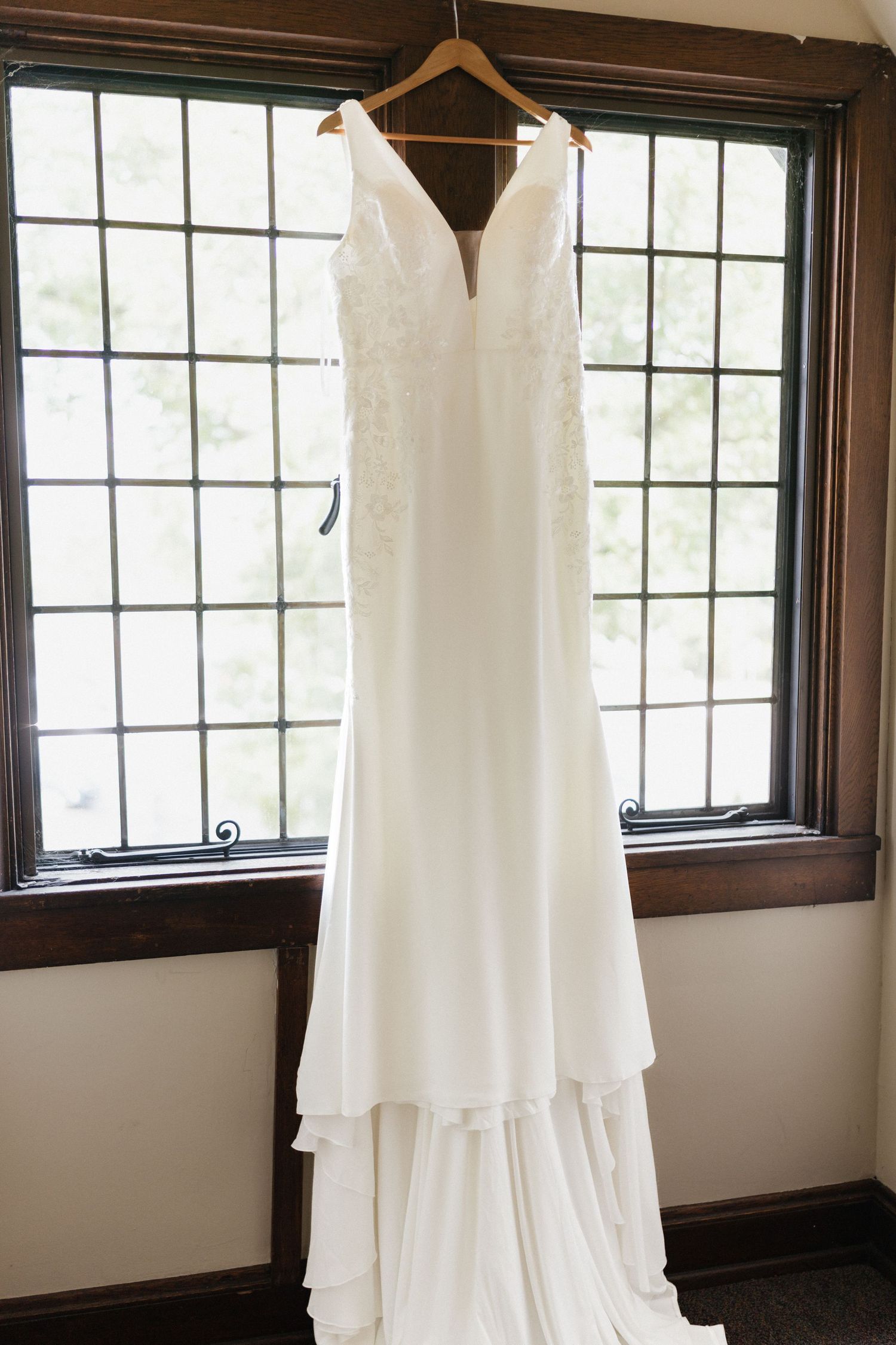 Three elegant white wedding dresses hanging on wooden hangers in a sunlit room with dark-framed windows.