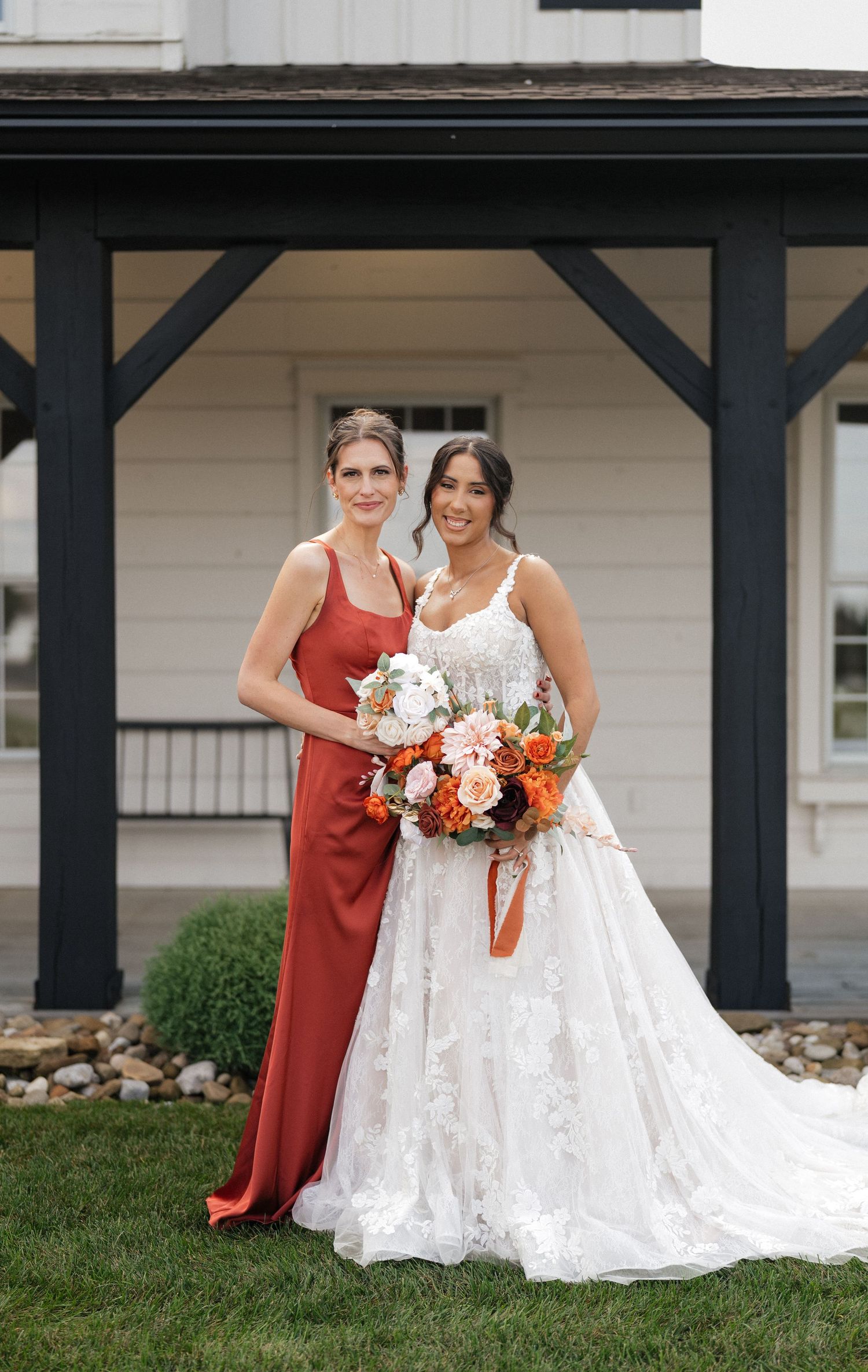 A bridesmaid in rust-colored dress poses with bride holding vibrant orange and white bouquet under covered porch.