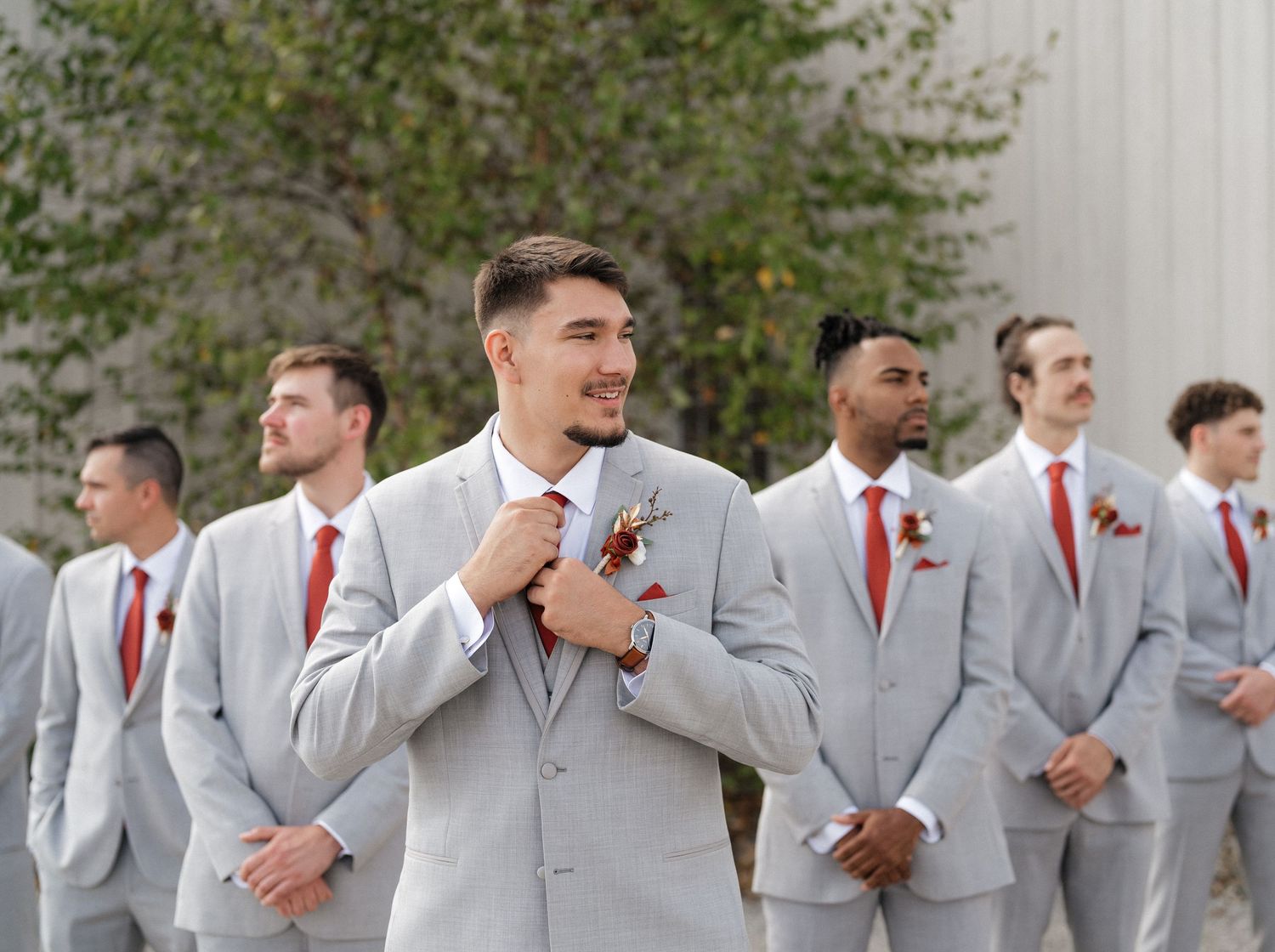 Groomsmen in matching light gray suits with red ties stand together outdoors.