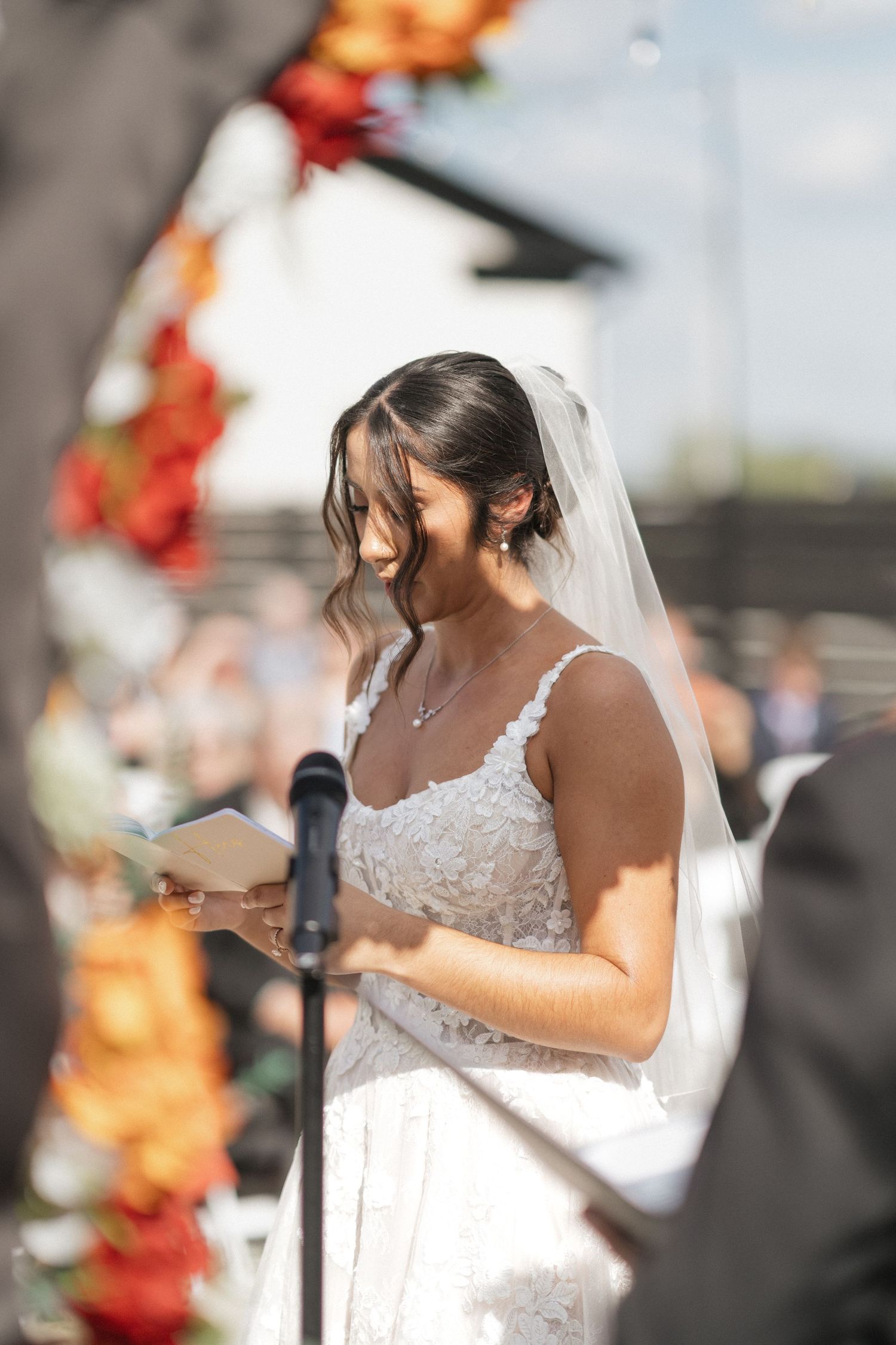 Person in white lace wedding dress reading vows at outdoor ceremony with orange floral arch.