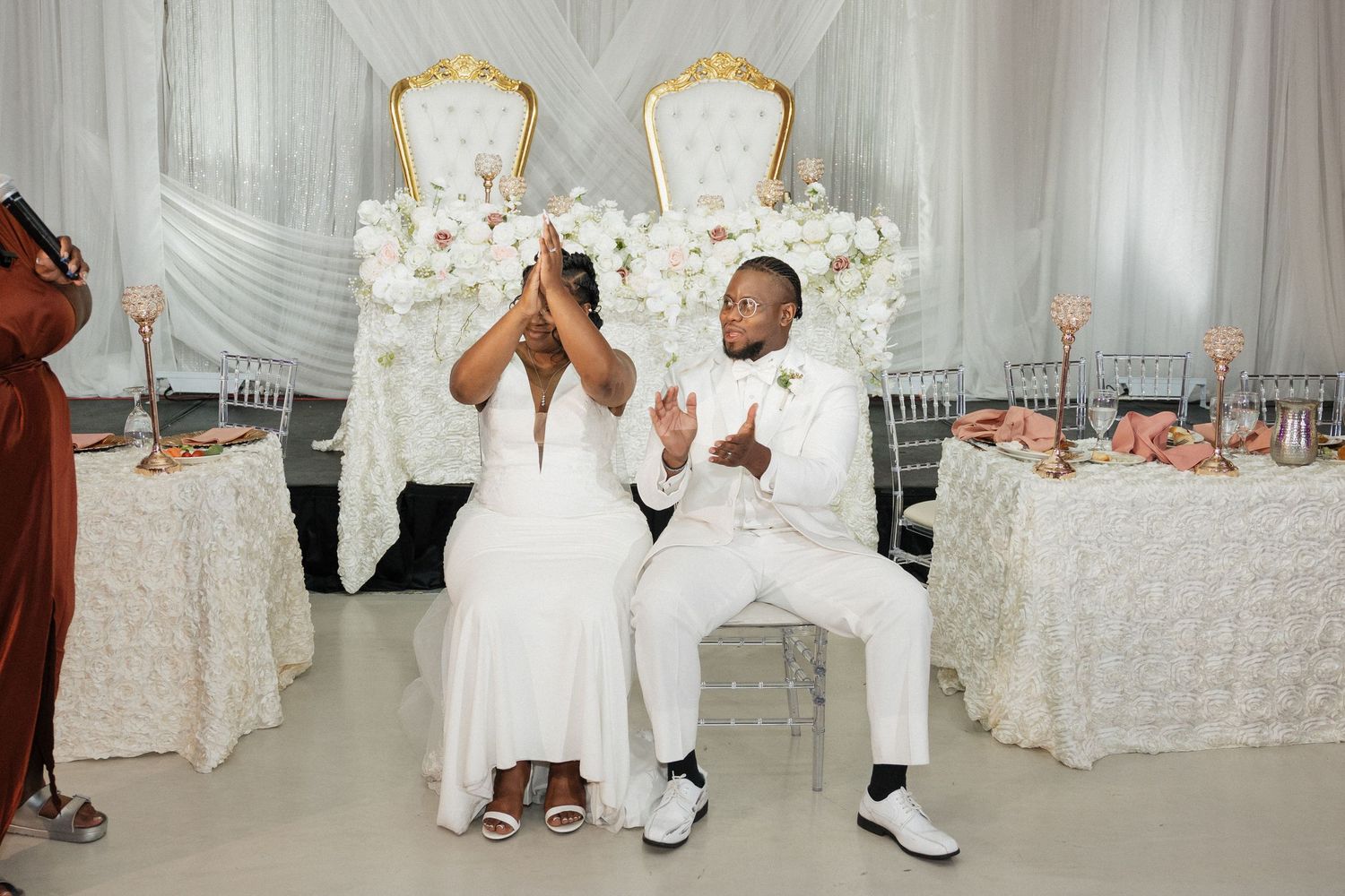 Couple sits on ornate white chairs at decorated wedding reception table.