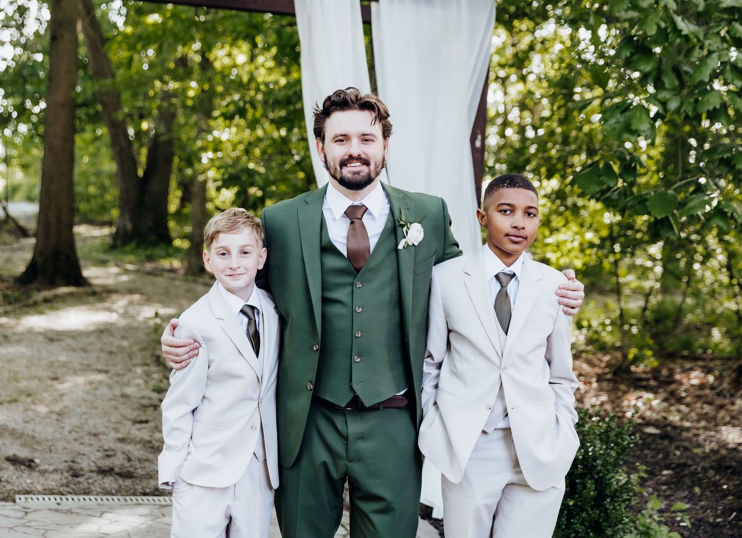 Ring bearers in crisp white suits stand with groomsman on stone patio outdoors.