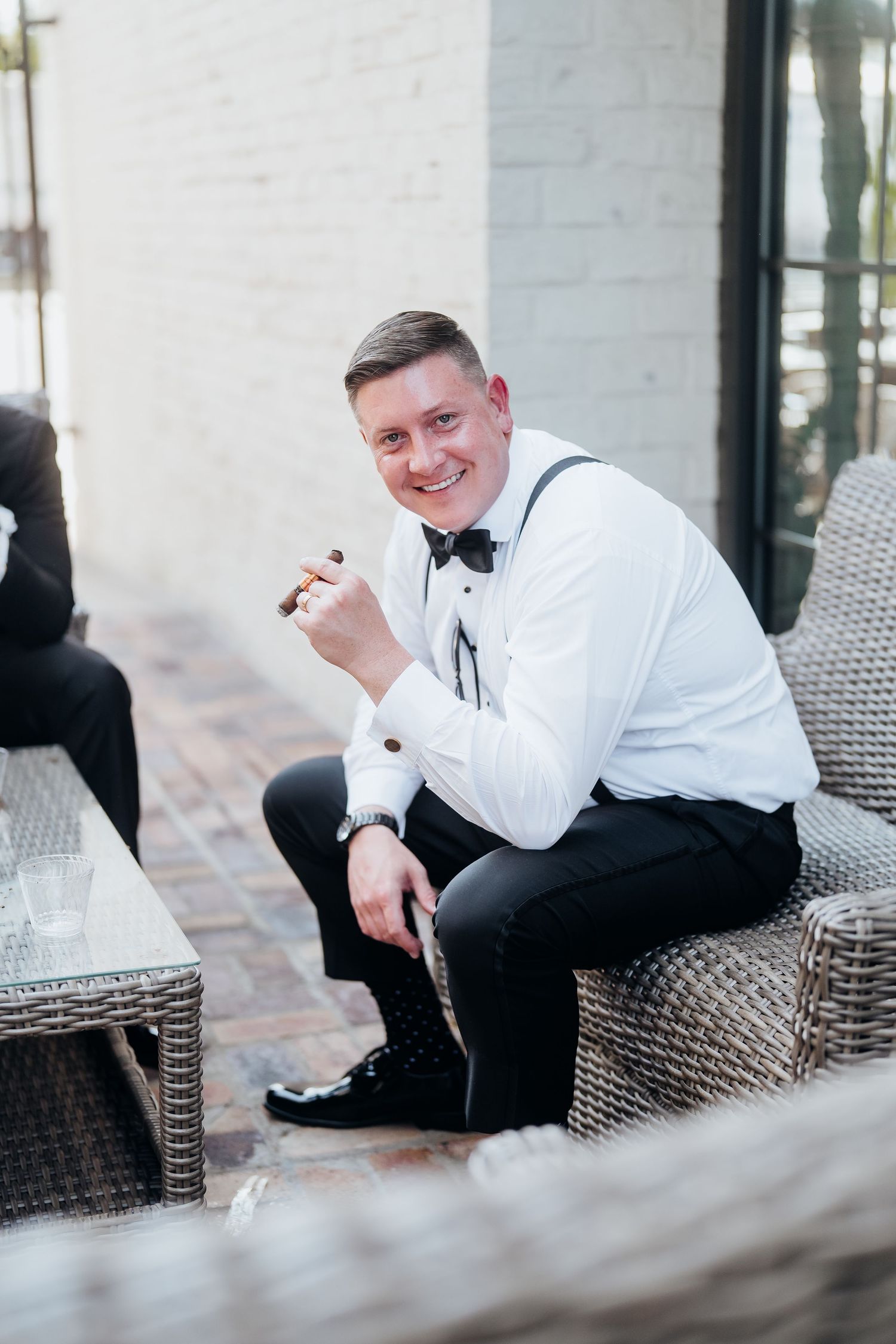 Groom in classic black and white attire poses casually on outdoor wicker furniture.