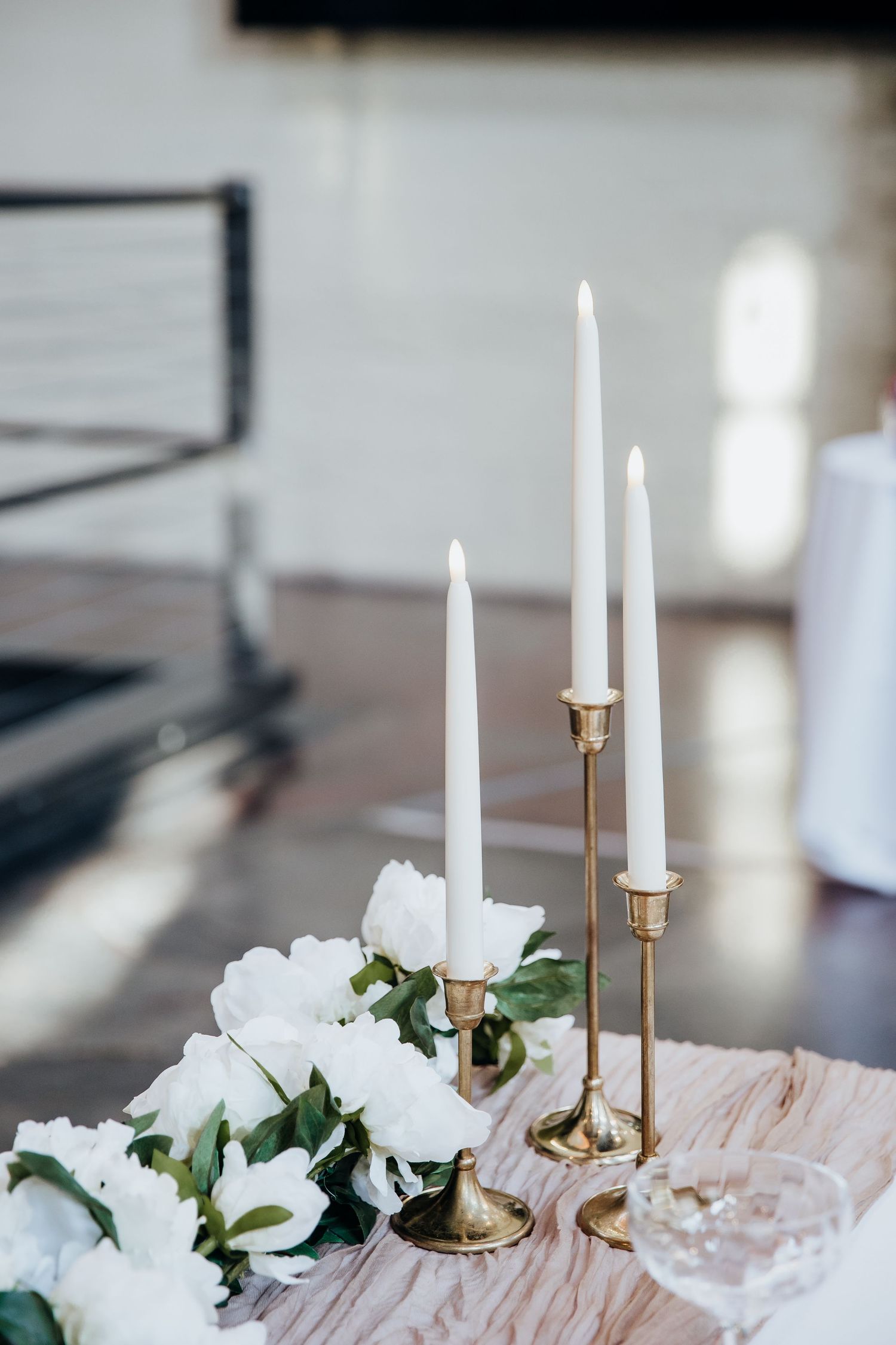 White taper candles in gold holders with white floral arrangements on blush table linens.