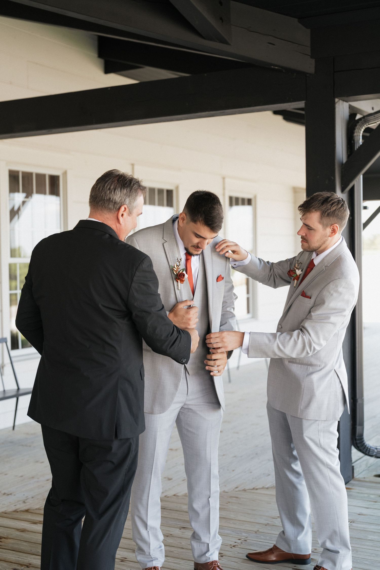Groomsmen help adjust ties and suits before a wedding ceremony in a light-filled venue.