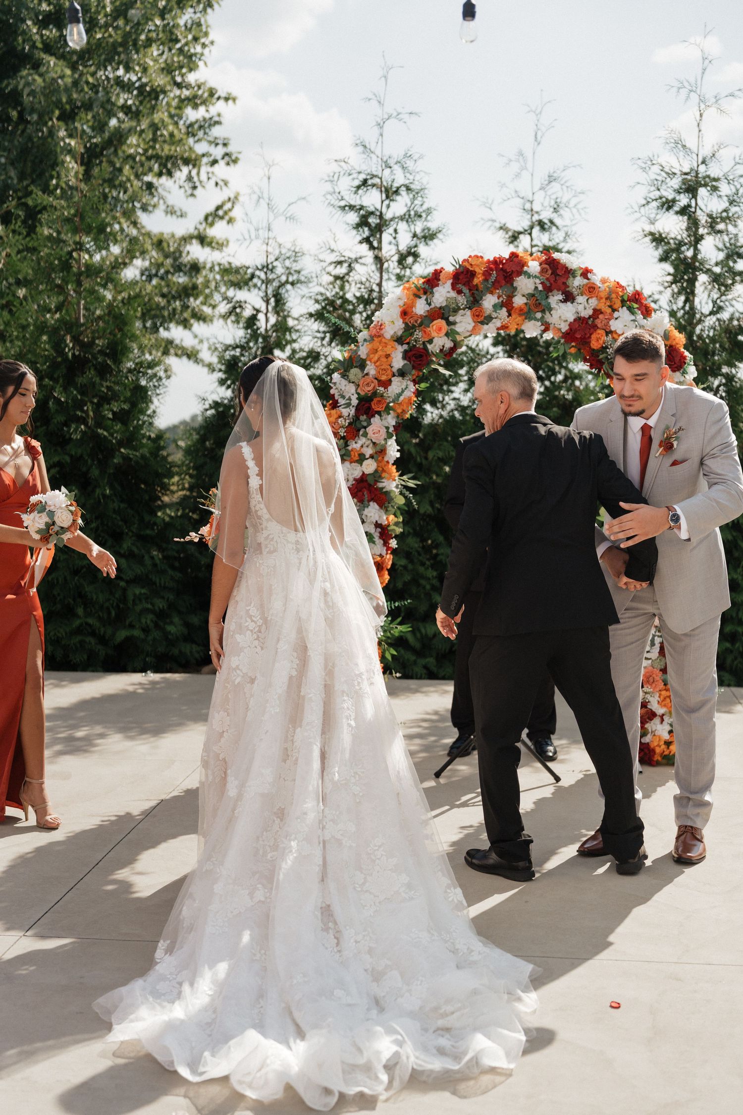 Wedding ceremony under a floral arch decorated with orange and white flowers on a sunny outdoor patio.