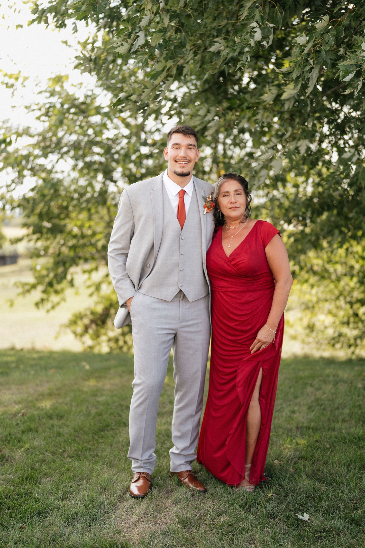 Wedding couple in grey suit and red dress posing together on grassy lawn with trees.