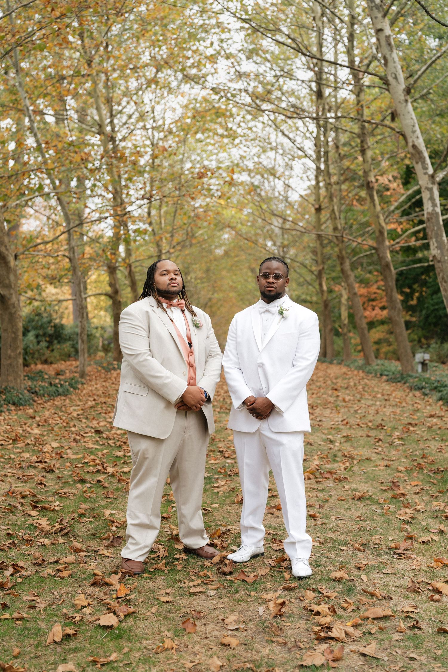 Two people in light colored suits stand together on an autumn path covered in fallen leaves.
