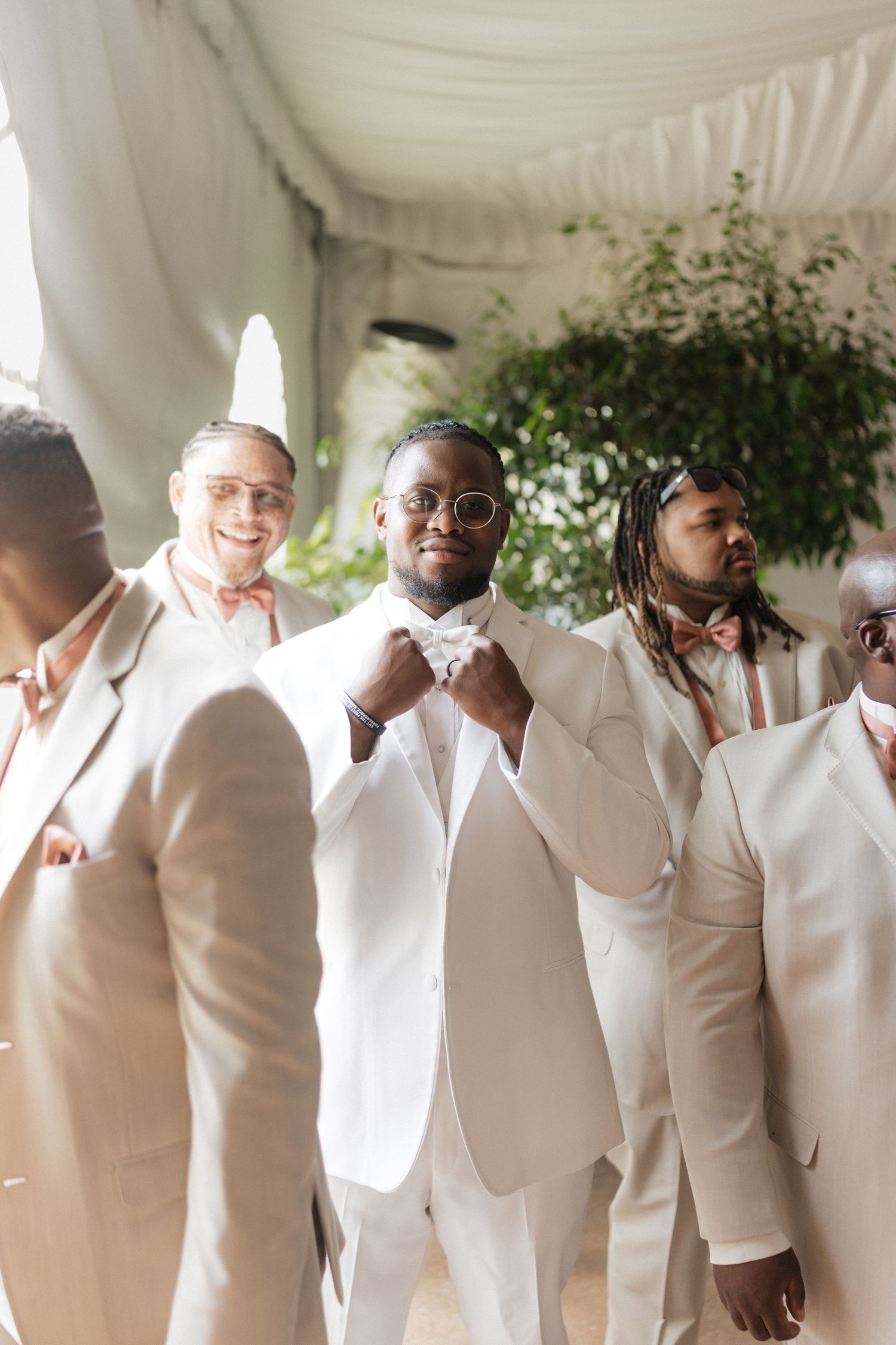 Groomsmen in crisp white suits and coral bow ties adjust their attire while standing under a tent.