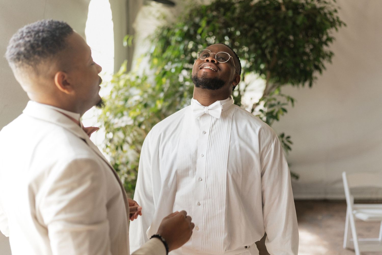 Two people in white attire having an animated conversation in a bright indoor space with greenery in the background.